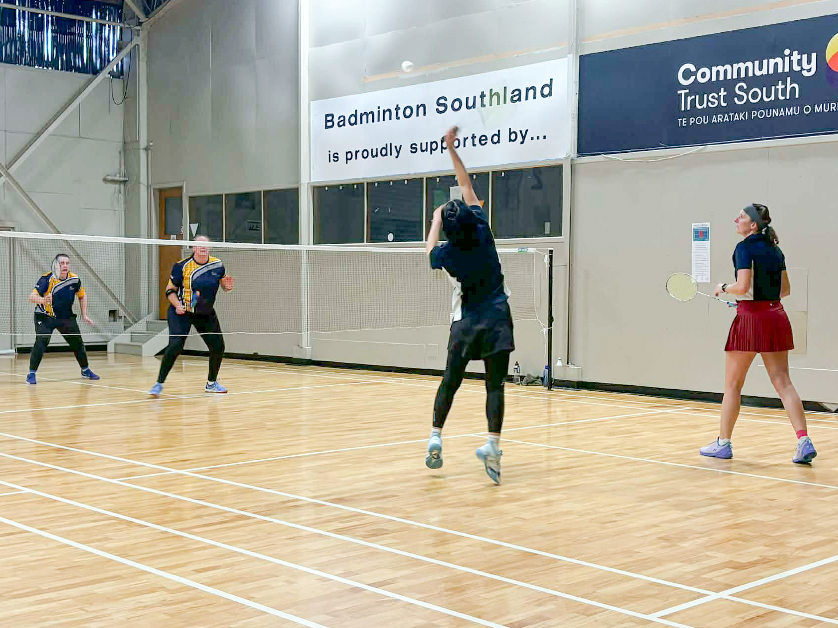 People playing badminton on an indoor court with wooden flooring and white walls. One person is mid-air hitting the shuttlecock, while others watch nearby.