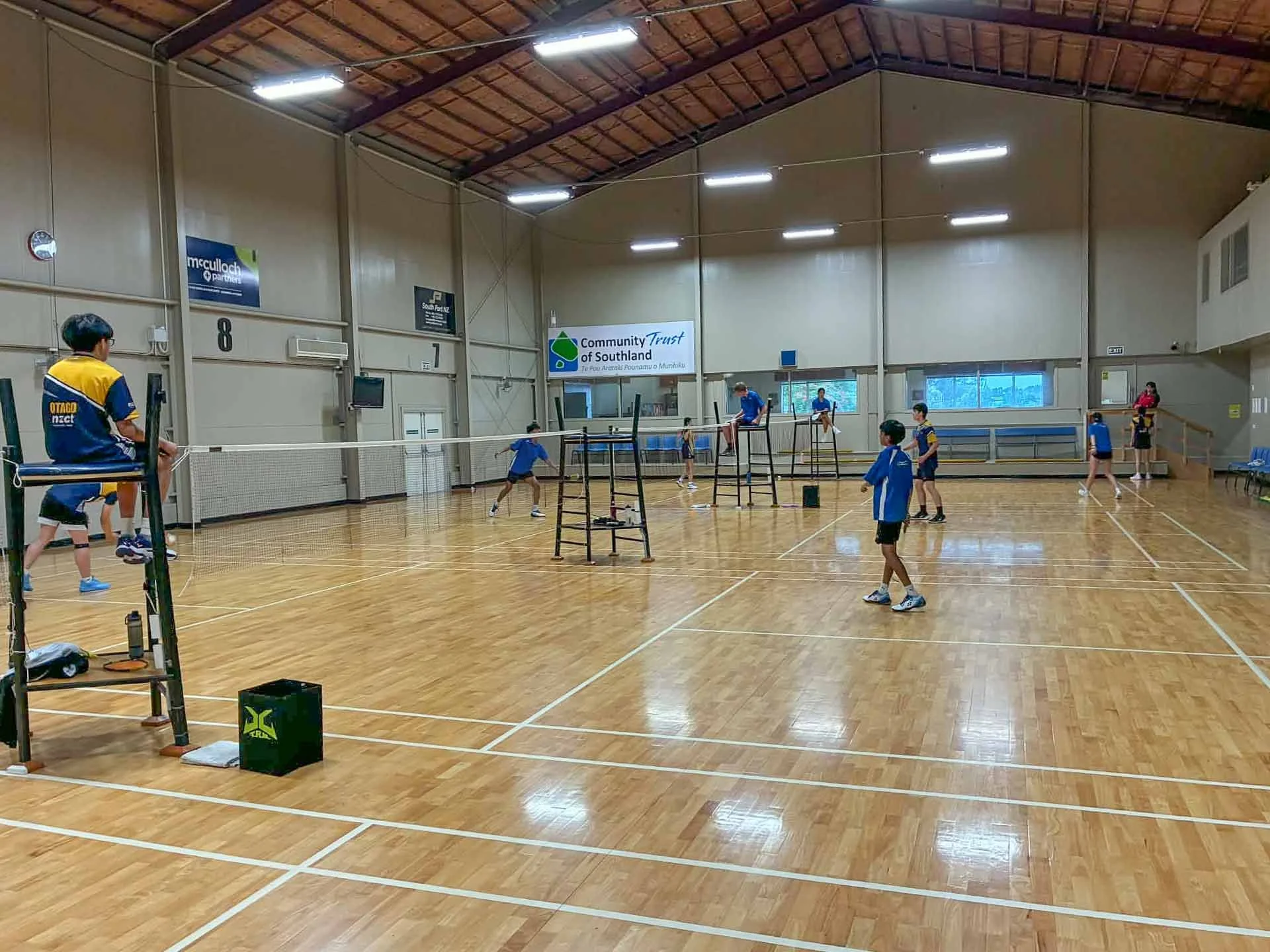 Indoor gymnasium with children playing volleyball, some children on the court and some in the background, there are banners and a clock on the walls.