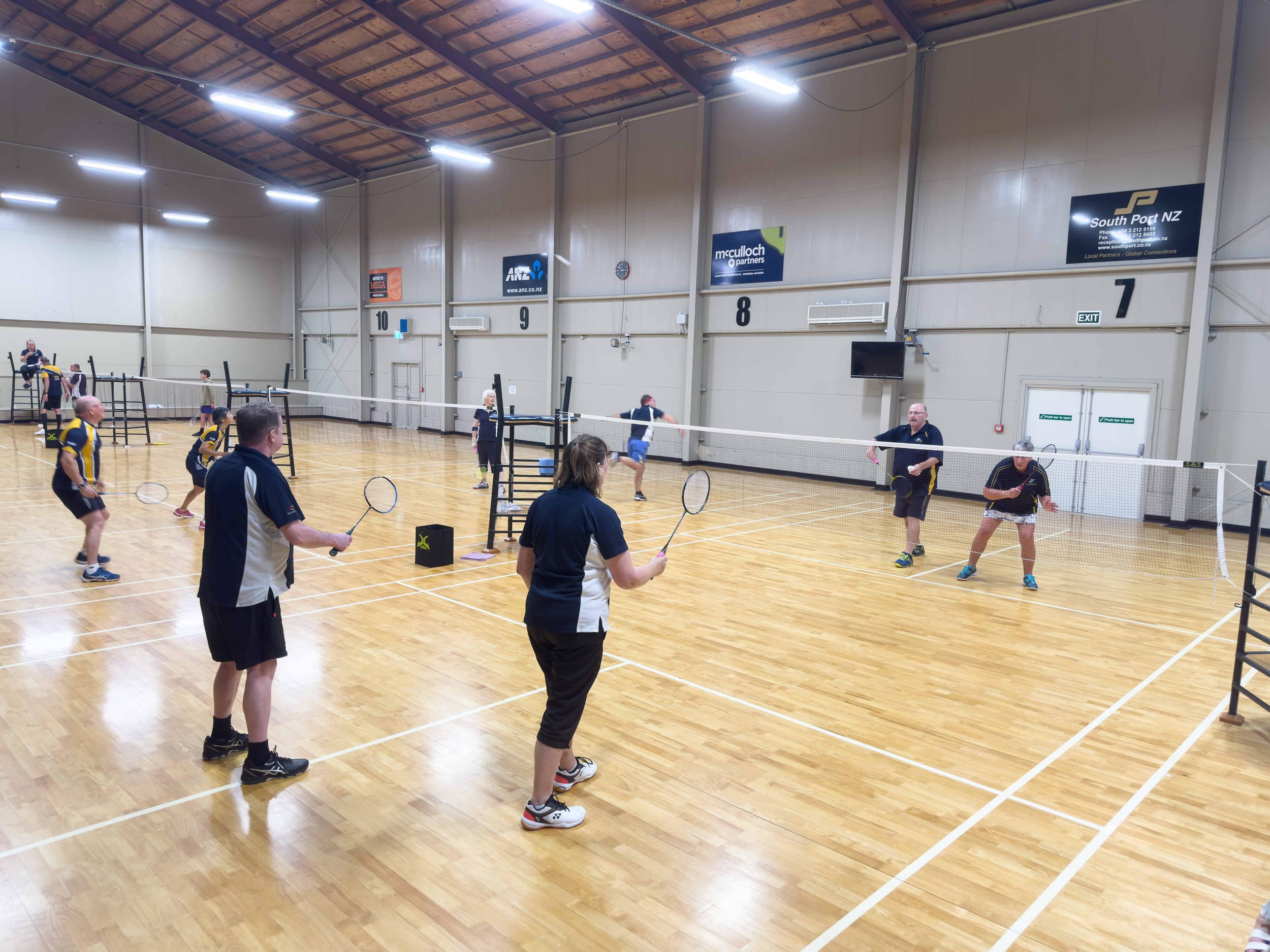 People playing badminton in an indoor sports hall with wooden flooring and high ceiling.