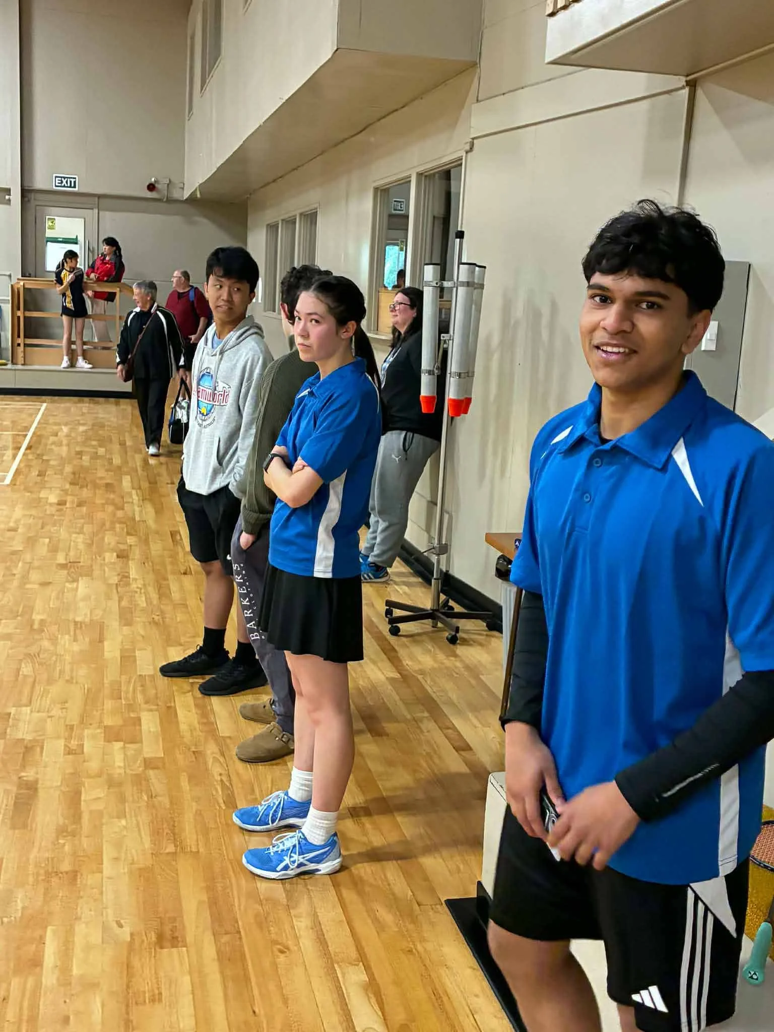 Young athletes standing in a gymnasium, lining up for an event or activity, with some spectators and other participants in the background.
