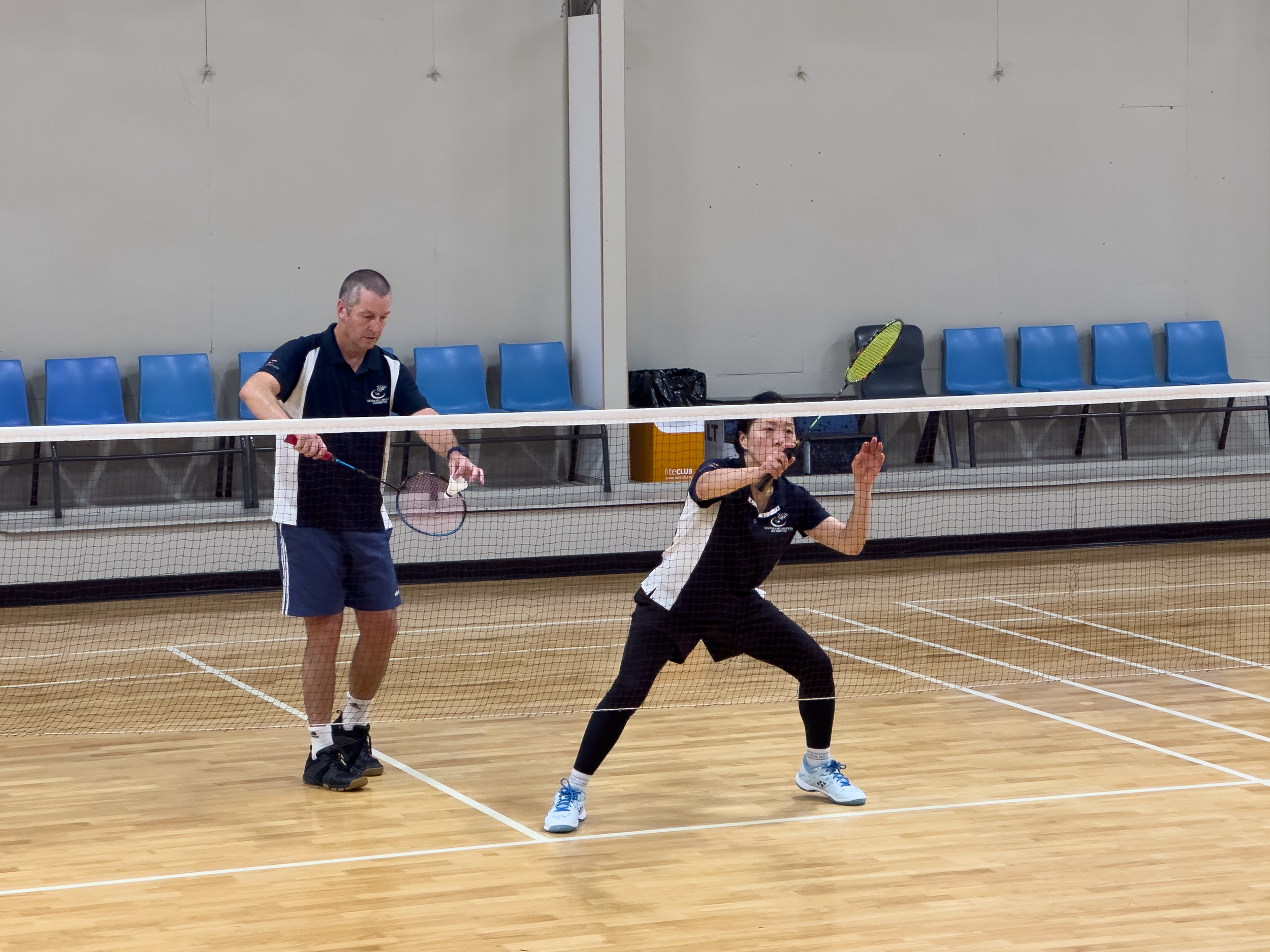 Two people playing badminton on an indoor court with a wooden floor and blue chairs in the background. The player in the foreground is a woman preparing to hit the shuttlecock, and the man in the background is holding a racket.