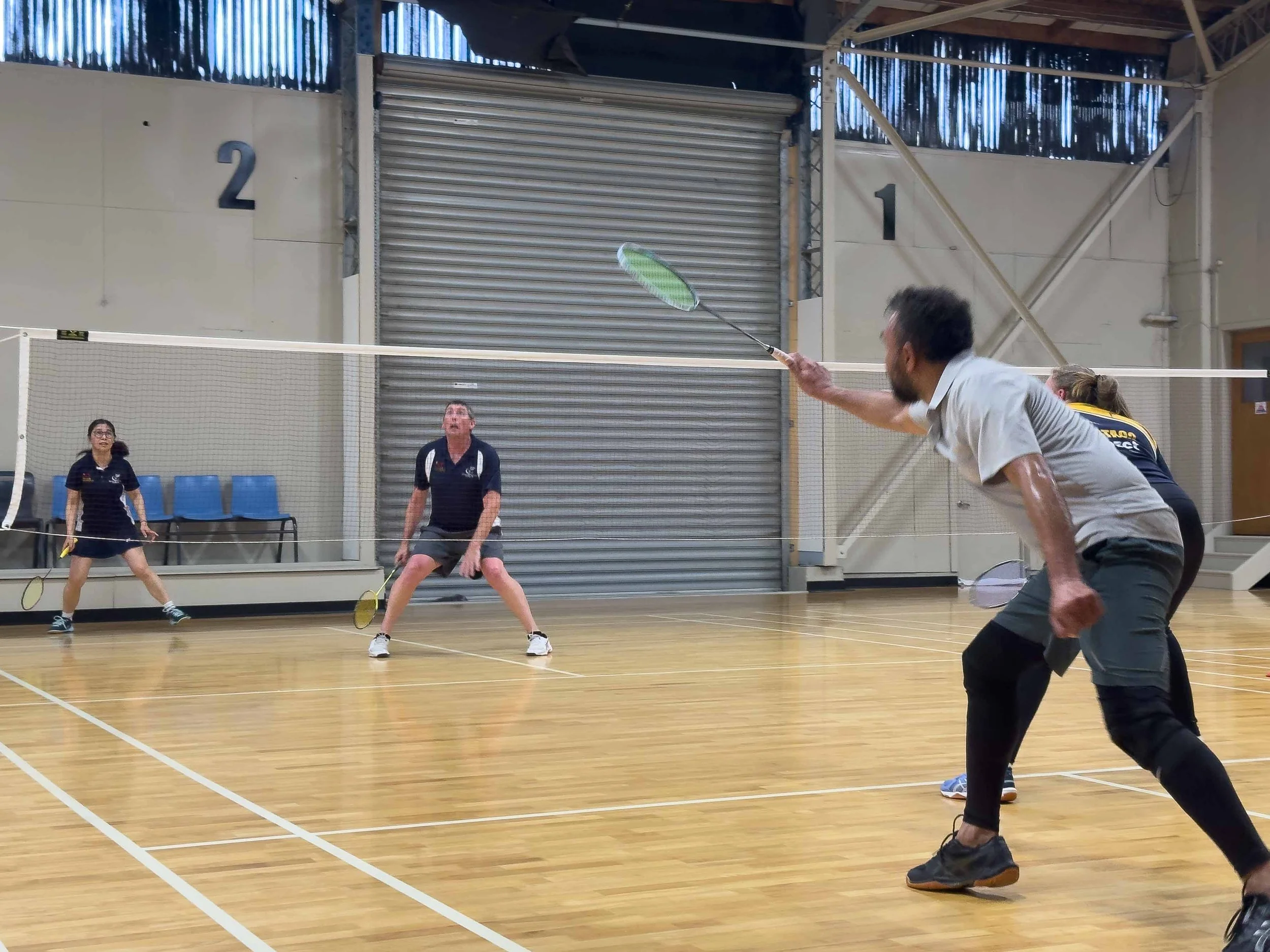 People playing badminton indoors on a wooden court, with two players on each side of the net, preparing to hit the shuttlecock.