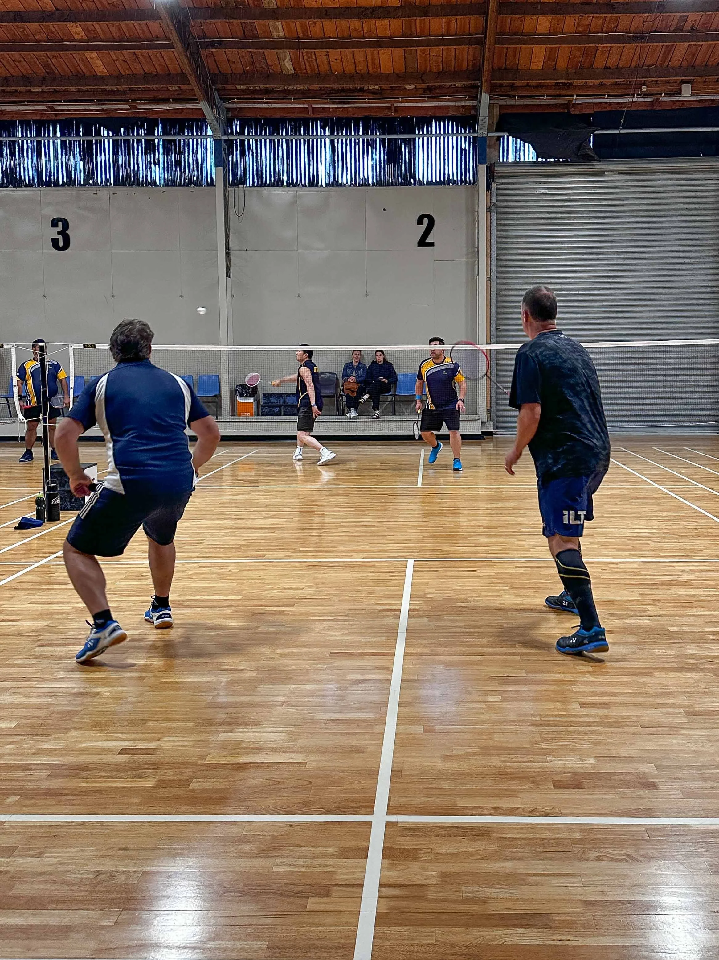 Indoor badminton game with players on a wooden court, some in action and some sitting on benches, with a partially open roller door in the background.