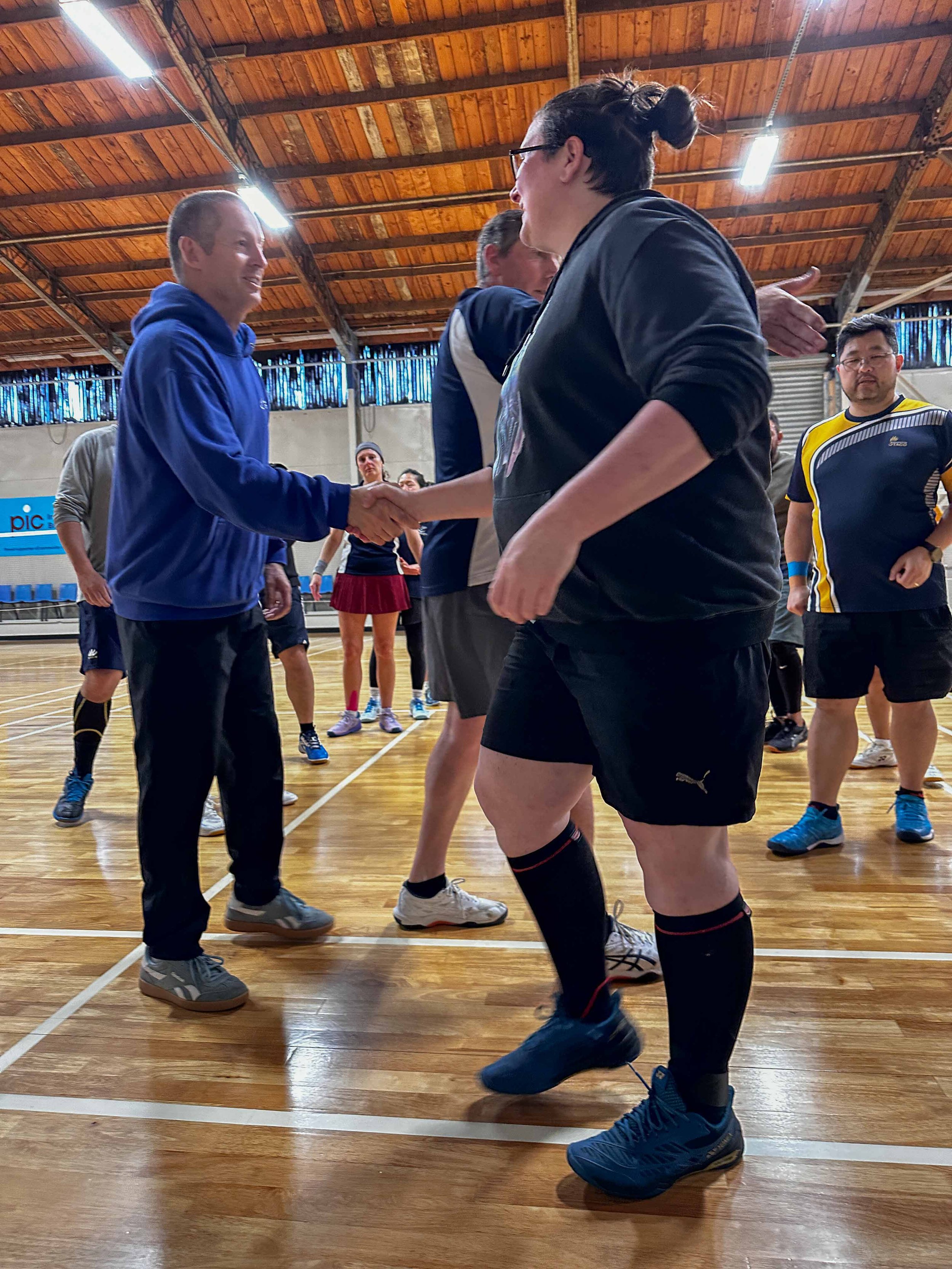 Group of people in sportswear inside a gymnasium, shaking hands, with sports equipment and a wooden ceiling in the background.