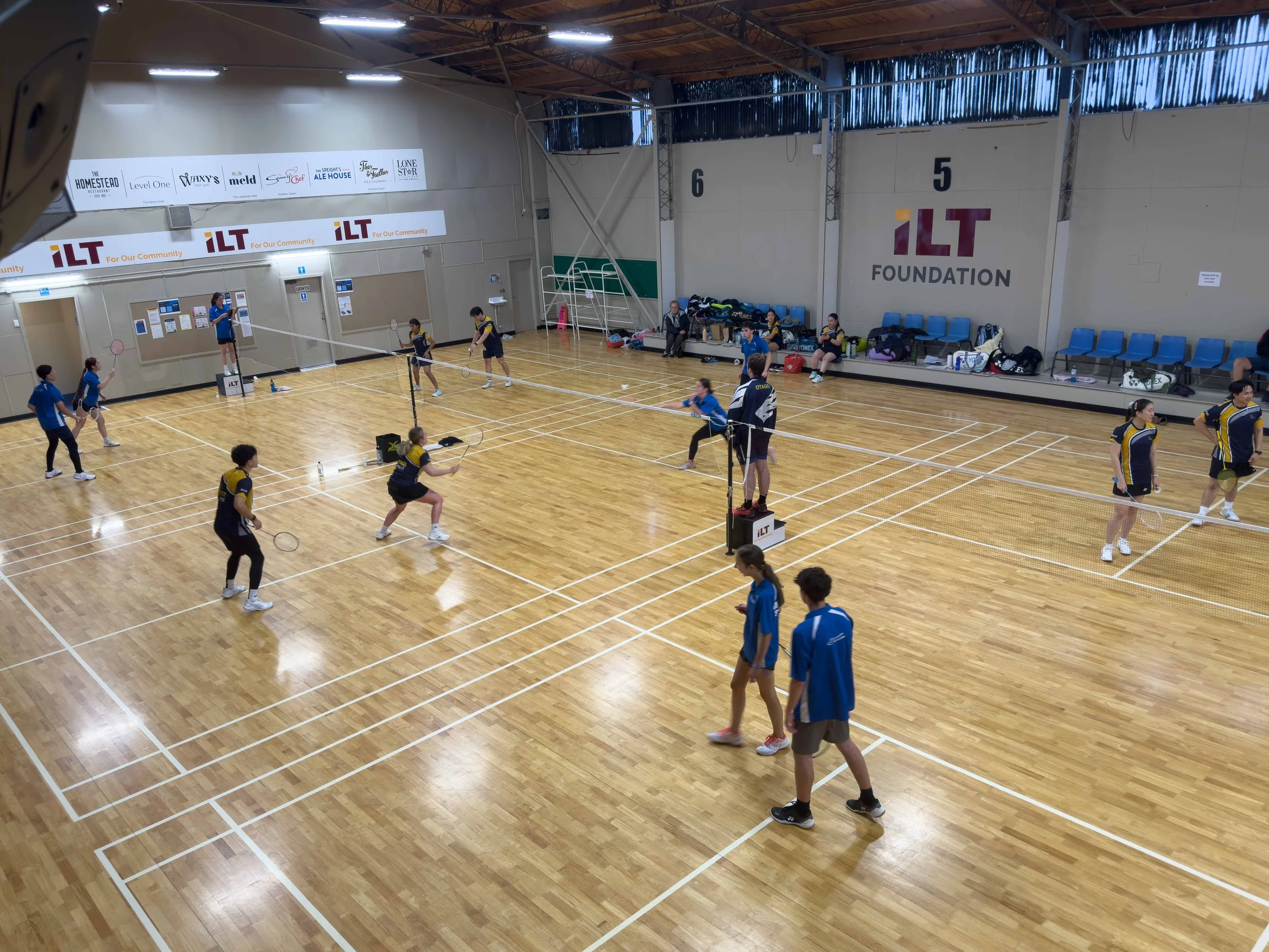 Indoor badminton court with players engaging in practice or game, b sitting on the sideline, and walls displaying ILT Foundation branding.