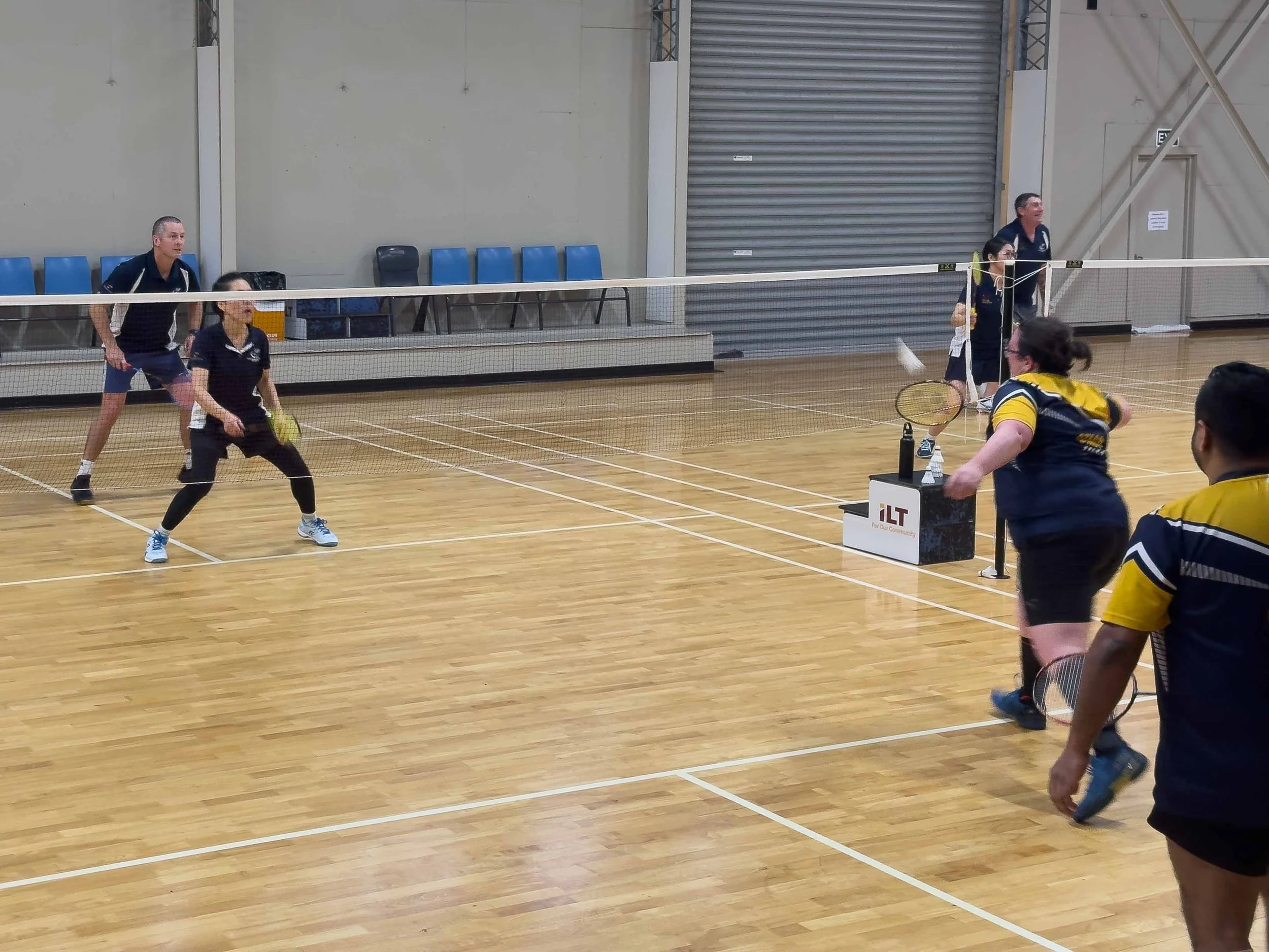 People playing badminton in an indoor gym with wood flooring, some playing on one side of the net and a few watching on the sidelines.