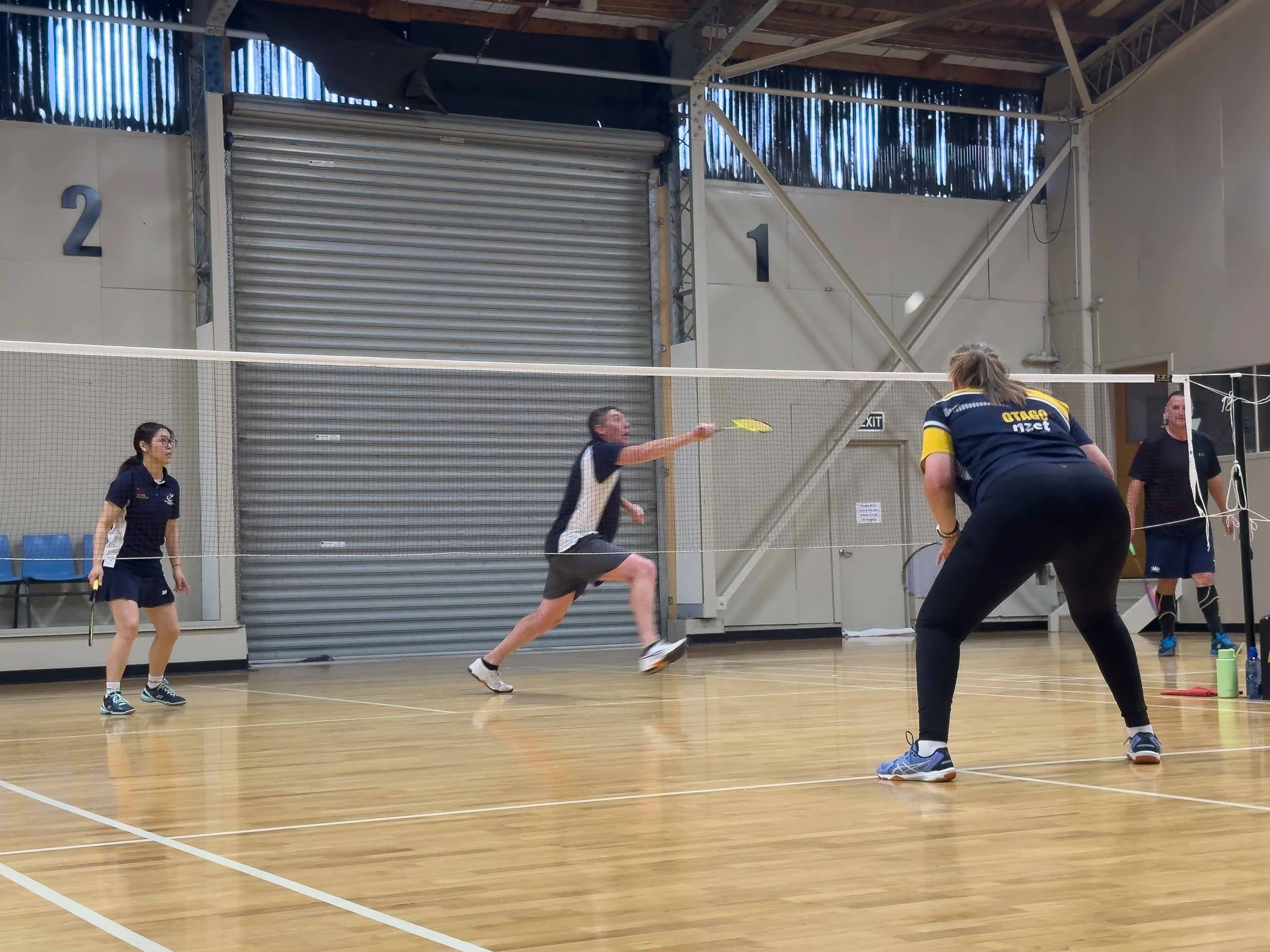 Four people playing badminton indoors, two women and two men. One woman is preparing to hit the shuttlecock, while the man in the center is lunging to return it. The other woman and man stand on the opposite side of the net, ready to play. The gym ha
