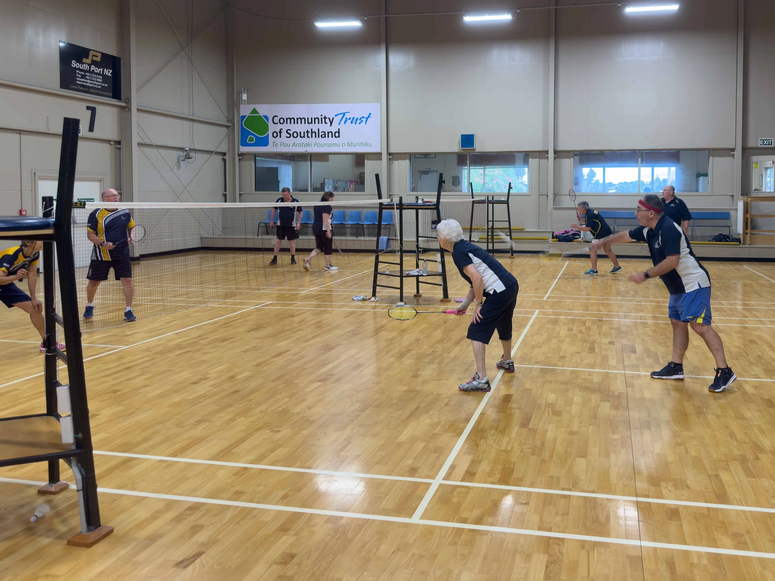 People playing pickleball indoors on a wooden court with a net, some holding paddles, with a sign behind them reading Community Trust of Southland.