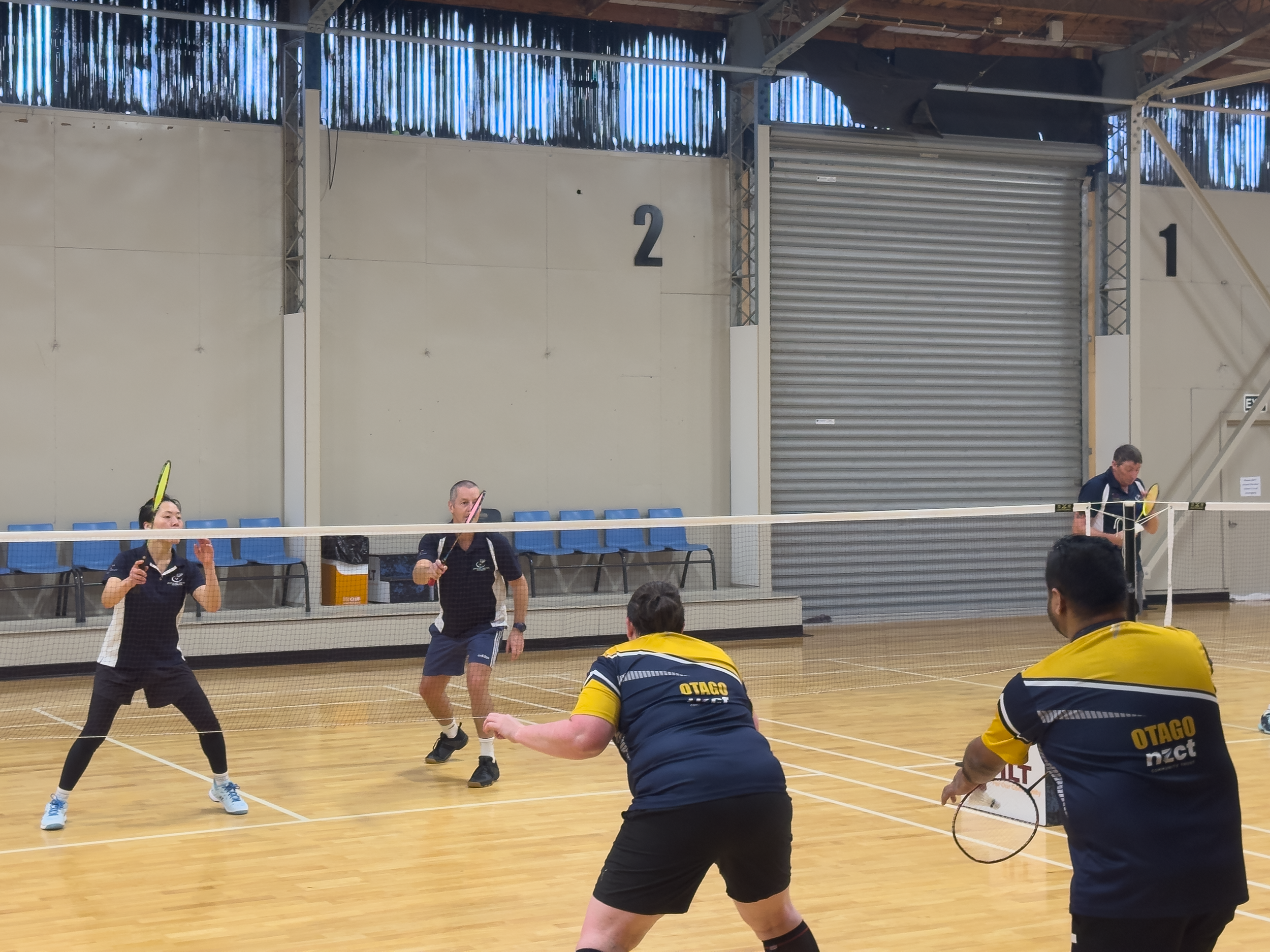 Two pairs of players engaging in a badminton match on an indoor court, with players in black and yellow jerseys, and a net in the center.