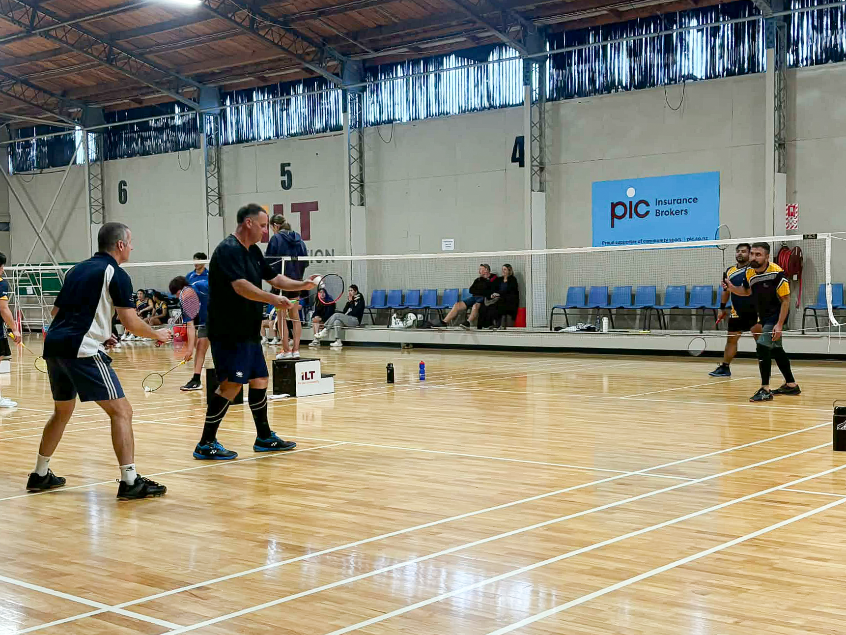 People playing badminton indoors on wooden court with benches and spectators watching in background.