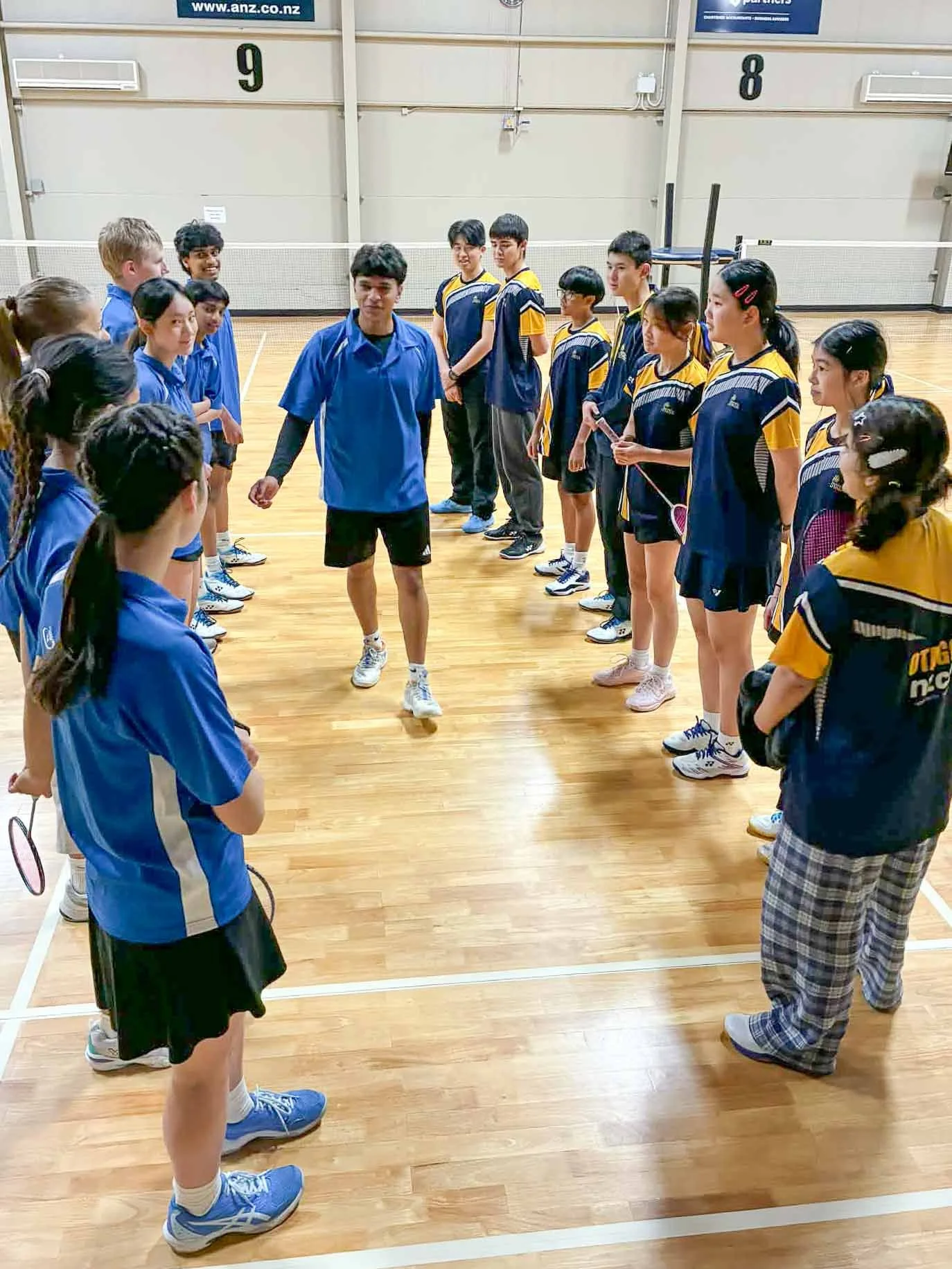 Group of children and a coach or teacher standing in a circle on a wooden indoor gym court, with badminton rackets, participating in a badminton activity or lesson.