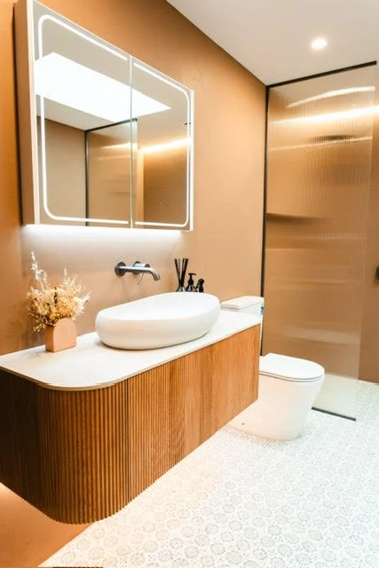 Modern bathroom with a white vessel sink, wooden vanity, illuminated mirror, and a beige wall with a glass shower enclosure.