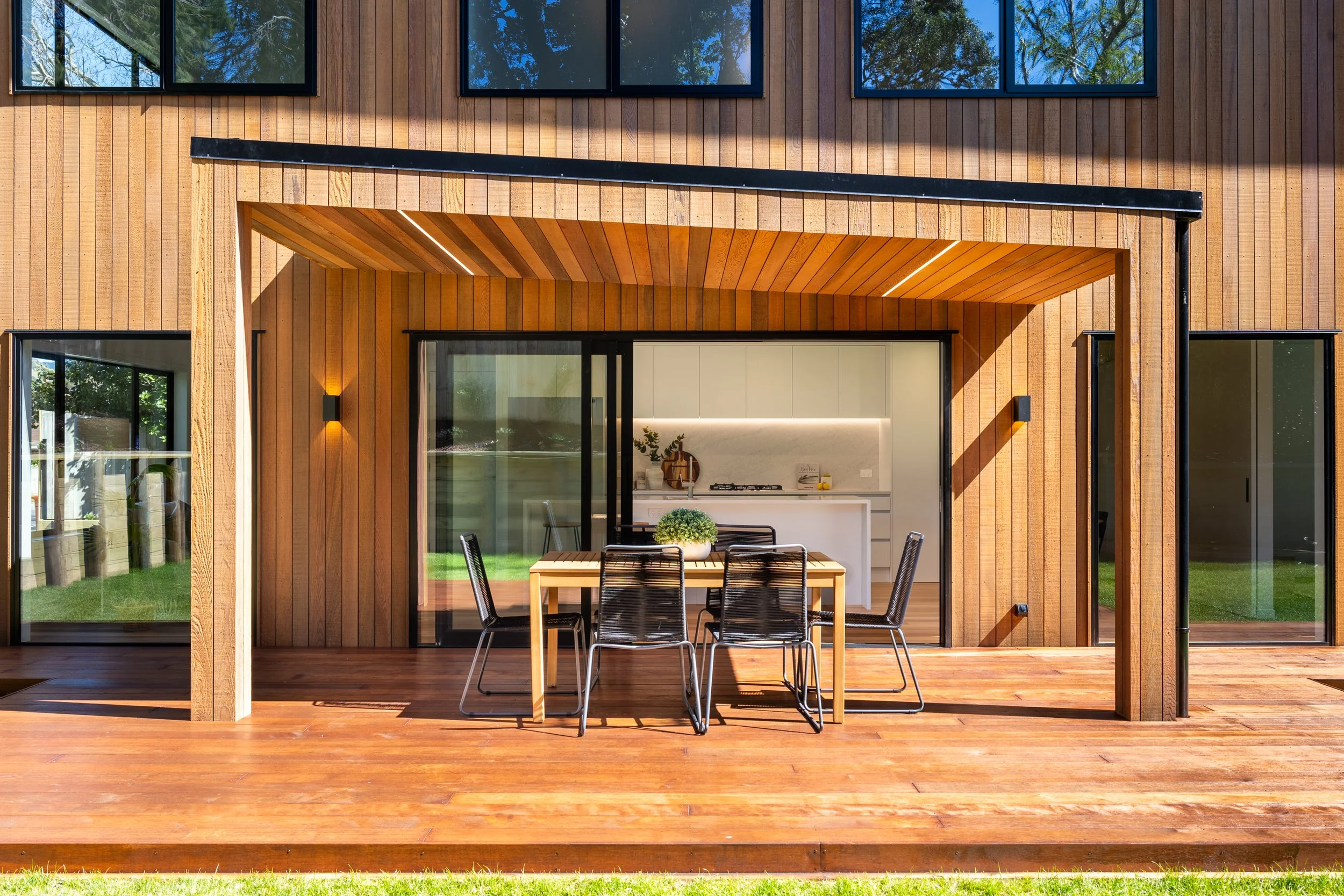 A modern house exterior with a wooden deck, large glass sliding door, and outdoor dining area with a table and six black chairs. The house has vertical wooden siding and large windows reflecting green trees.