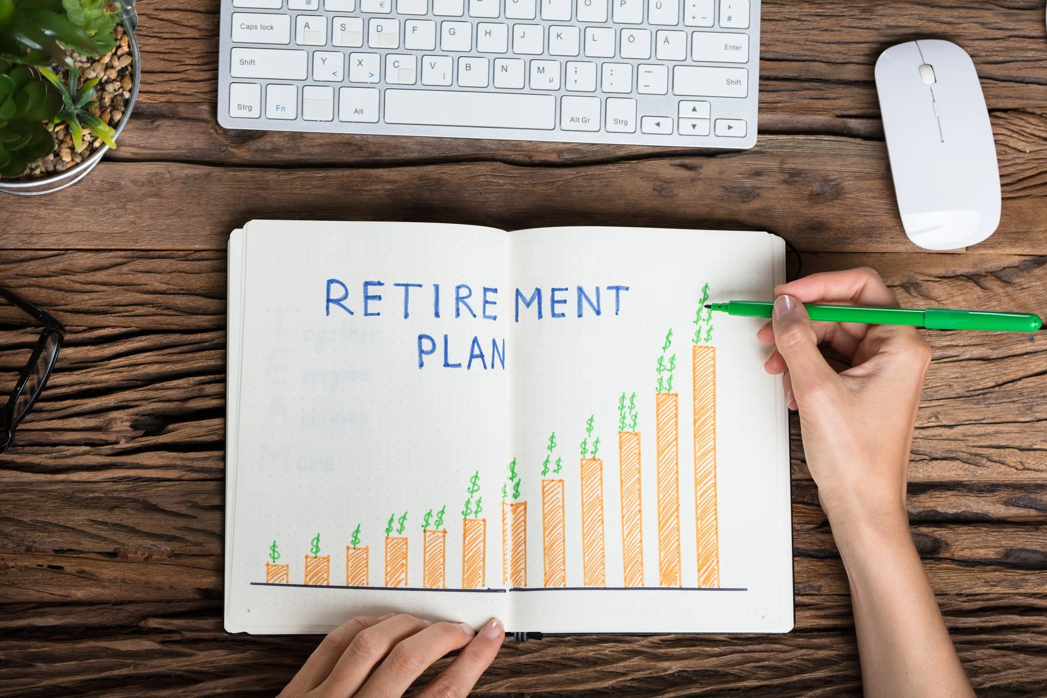 Open notebook with 'Retirement Plan' written on page and a bar graph with dollar signs, on a wooden desk with a white wireless keyboard, computer mouse, and a potted plant.