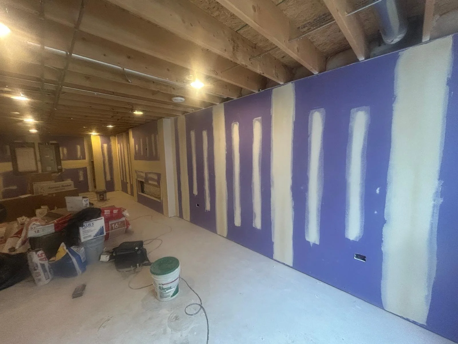 Interior of a room under construction or renovation, with drywall installed on the walls and ceiling, some drywall mudwork applied, and construction tools and materials scattered on the floor.