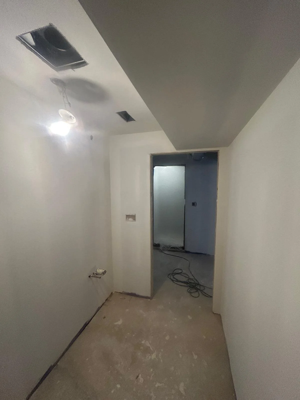 Empty room under construction with white walls, unfinished floor, and ceiling with vents and a light fixture. Open doorway leading to another room with exposed wiring.