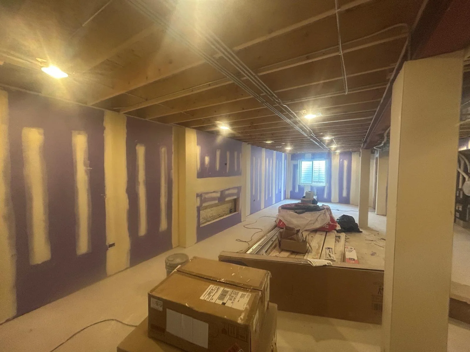 Interior of a room under construction with drywall and insulation partially installed, construction materials and tools scattered on the floor, a window at the far end of the room, and exposed ceiling framework.