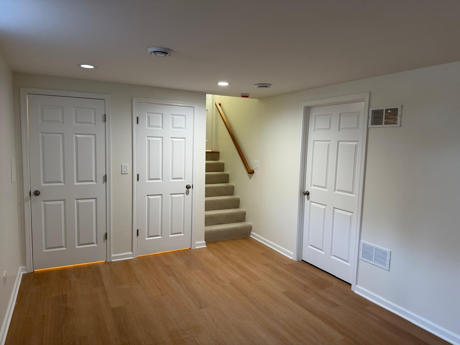 Empty room with wooden flooring, white walls, four white doors, a staircase with carpeted steps and a wooden handrail.