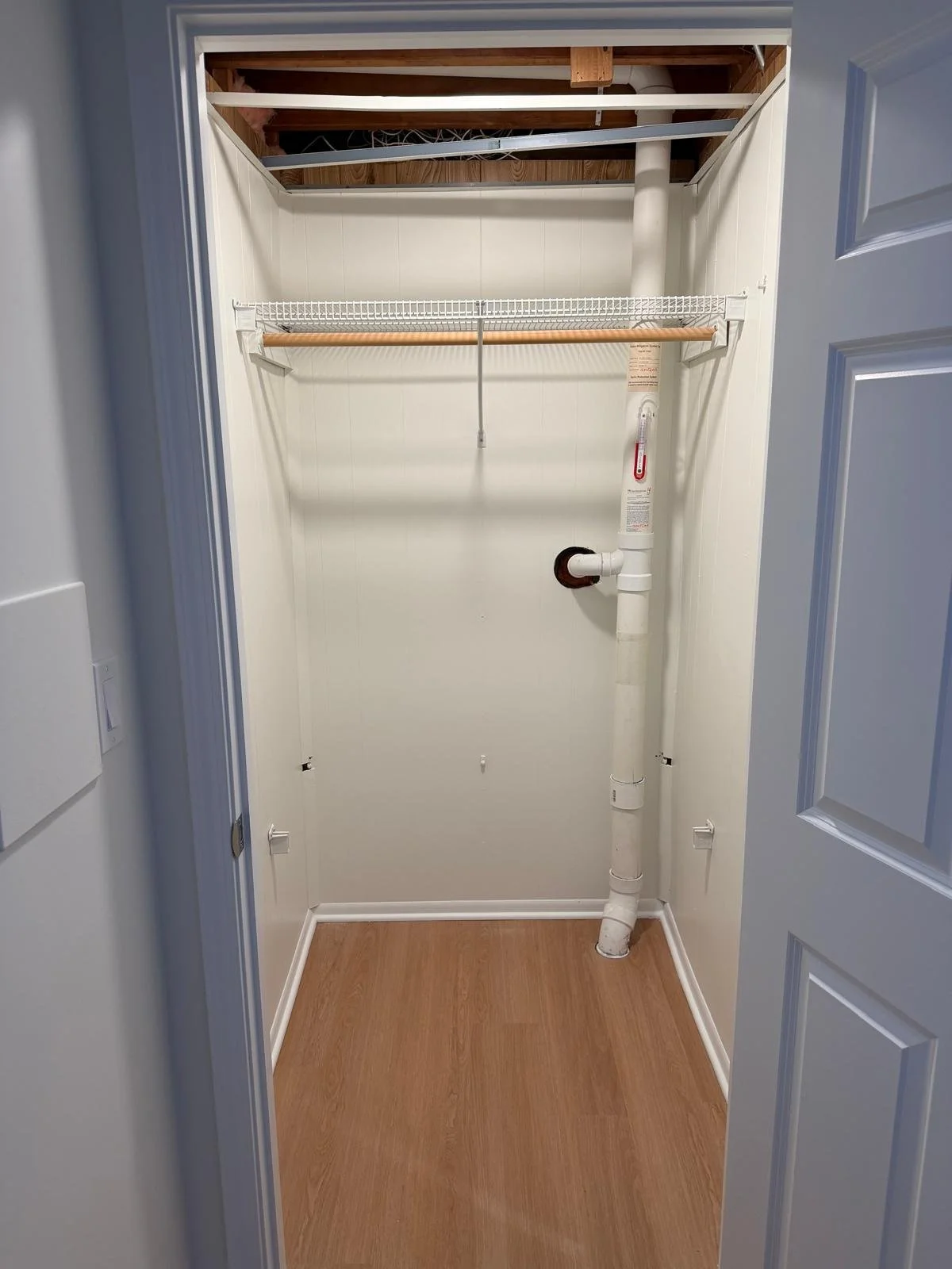 Empty closet with a white wire shelf, wooden hanging rod, exposed ceiling beams, and plumbing pipe on the right wall, with a wood floor and white walls.