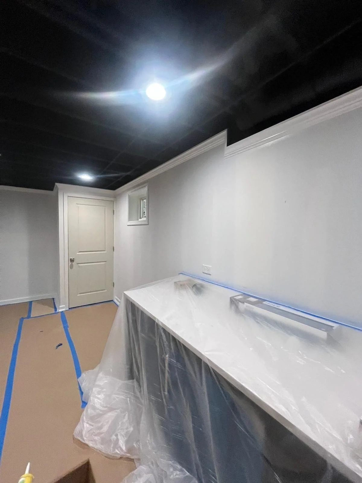 Room under construction with white walls, a door, a small window, and a black ceiling. Protective plastic covers and painter's tape are visible on the furniture and floor.