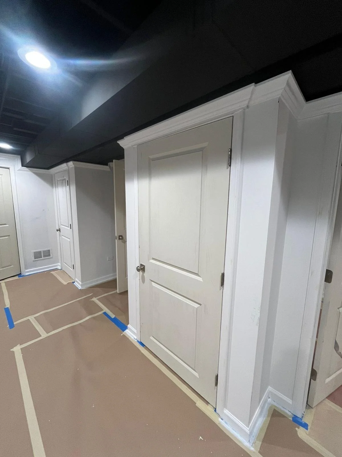 Interior view of a room with white walls and doors, some doors are slightly ajar, with brown paper and painter's tape on the floor, and a dark ceiling with recessed lighting.