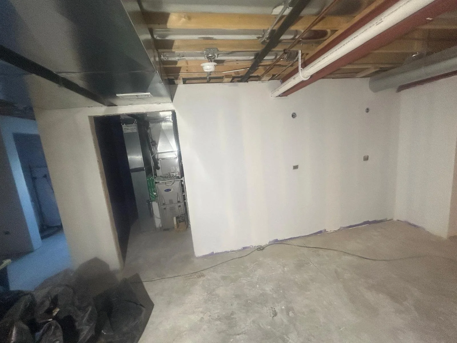 Undeveloped basement space with unfinished concrete floor, partially built wall with pipes and electrical outlets, open ceiling showing wooden beams, pipes, and ductwork, and a small utility room or kitchenette with plumbing fixtures.