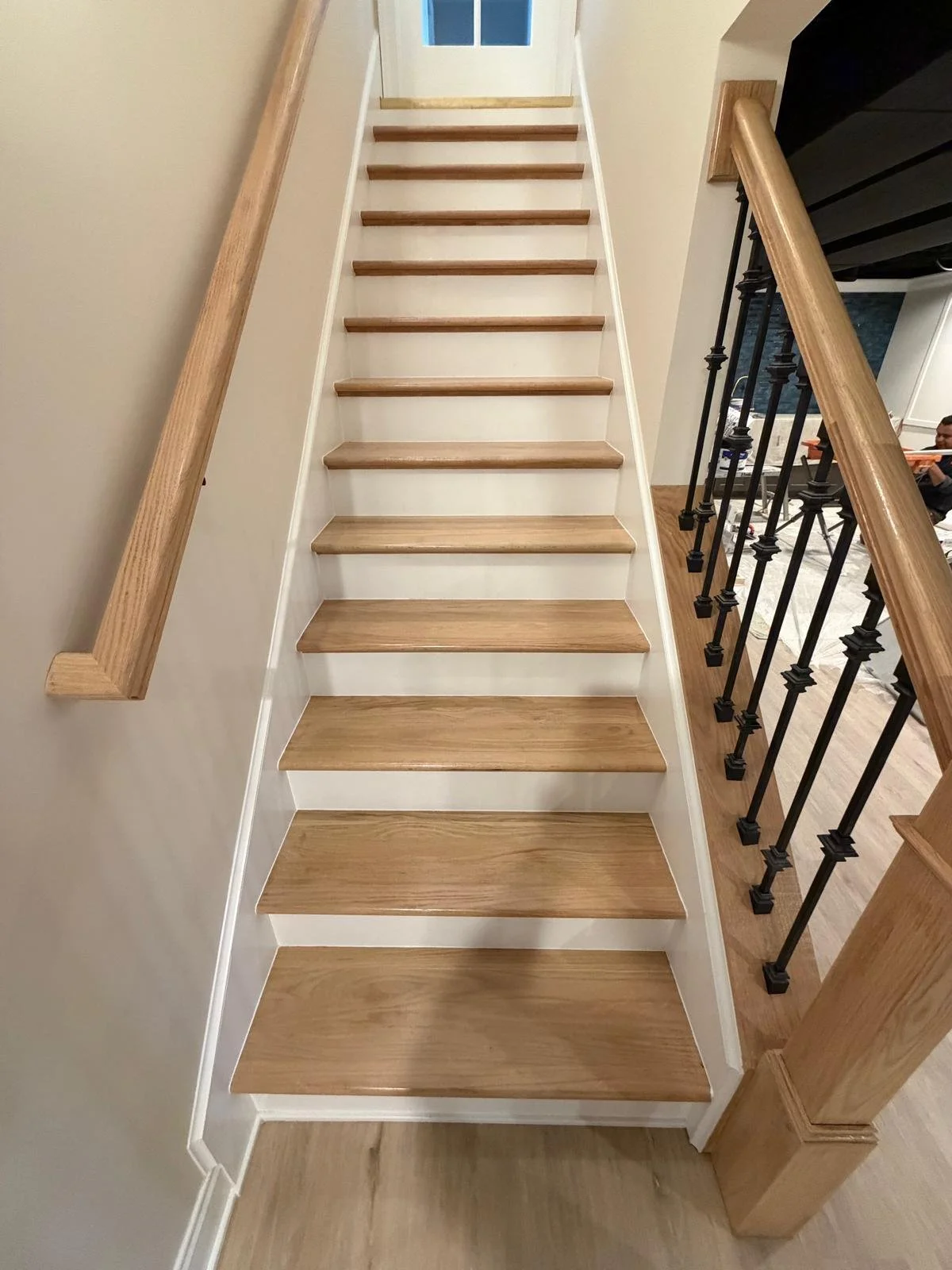 Interior view of a wooden staircase with light-colored wood steps, white risers, wooden handrails, and black metal balusters on the right side.