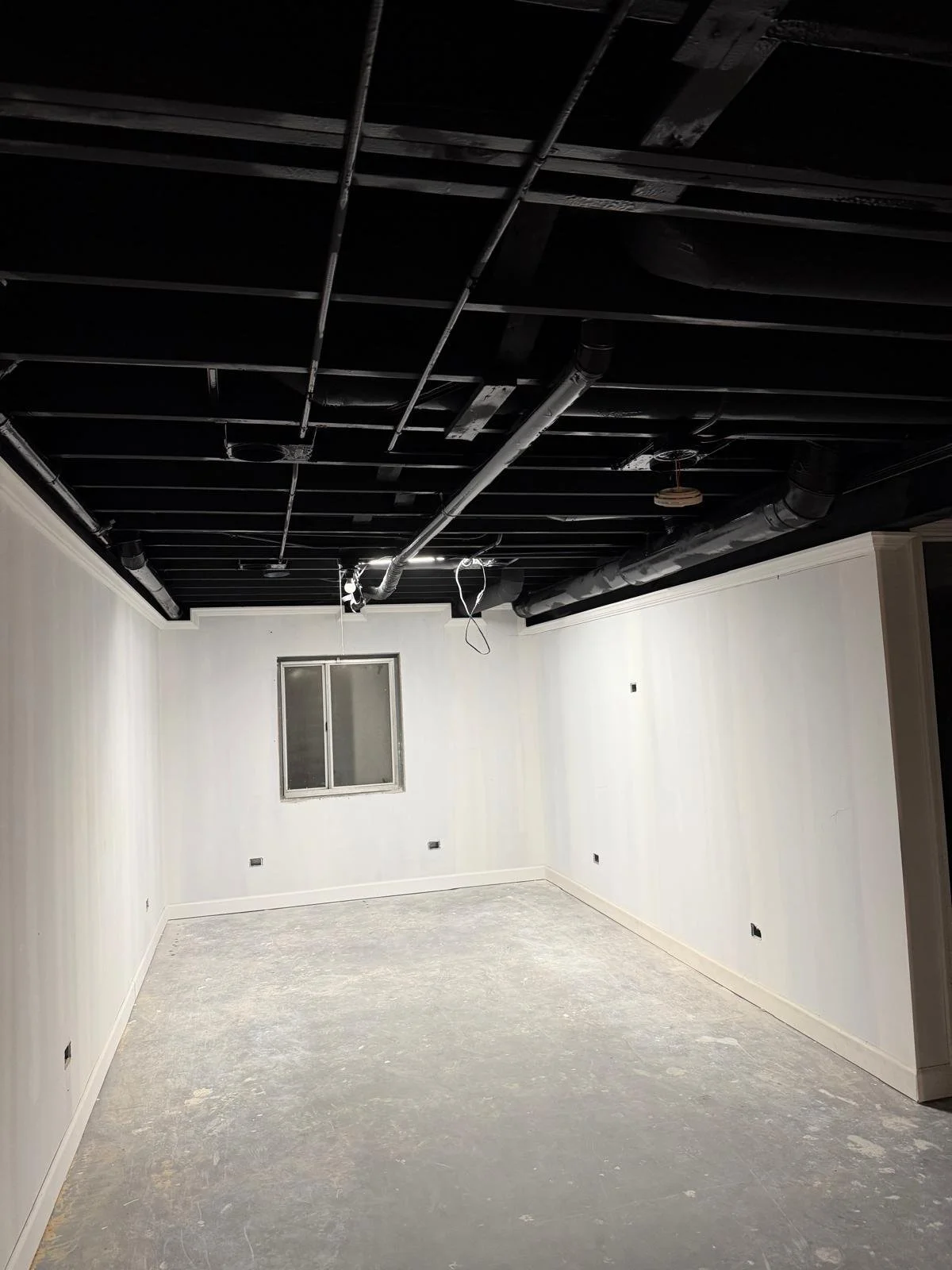 An unfinished room with white walls, a window, and a black ceiling with exposed pipes and wiring.