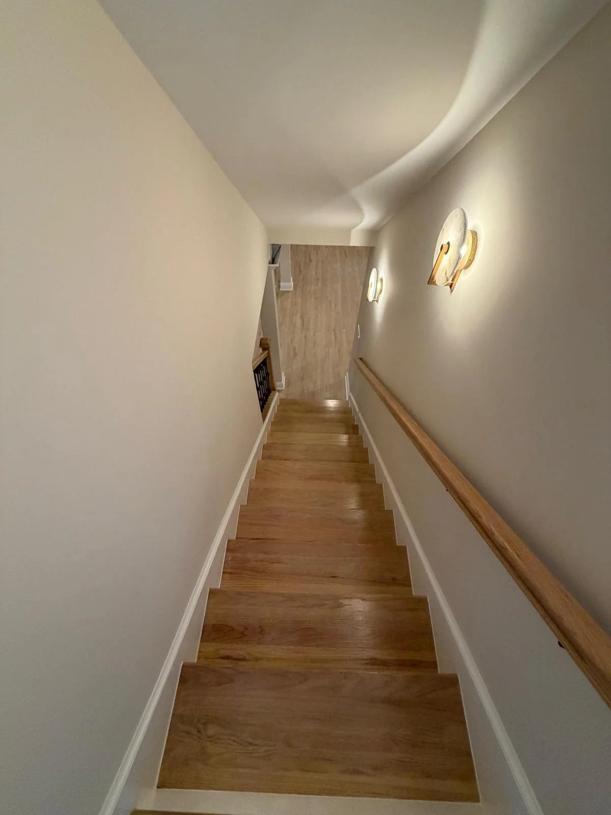 From the top of a wooden staircase, looking downwards into a hallway with wood flooring and light-colored walls, illuminated by wall-mounted light fixtures.