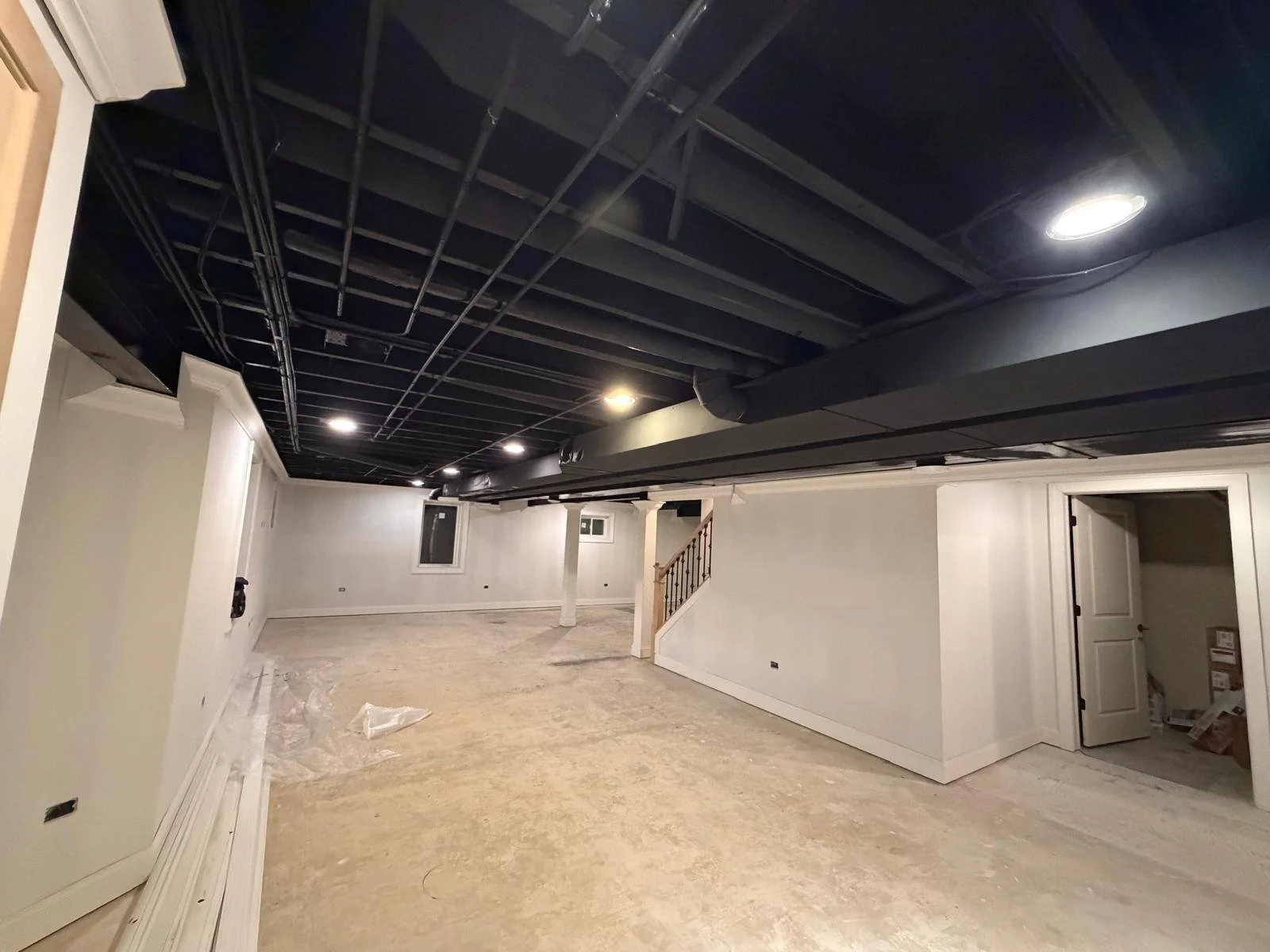 Under construction basement with black ceiling, white walls, and unfinished floor, with some wiring visible and a small stairway leading to an upper level.