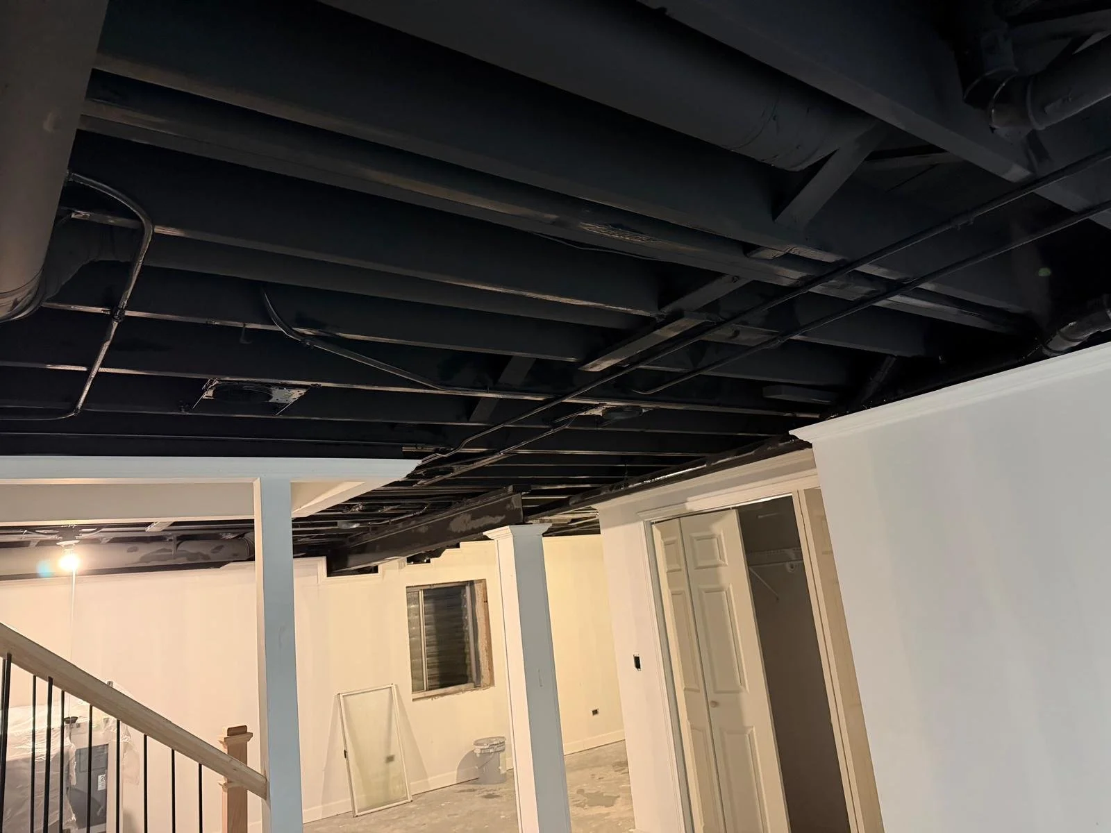 Ceiling with exposed black pipes and ducts in a building under construction or renovation, with part of a staircase and room in the background.
