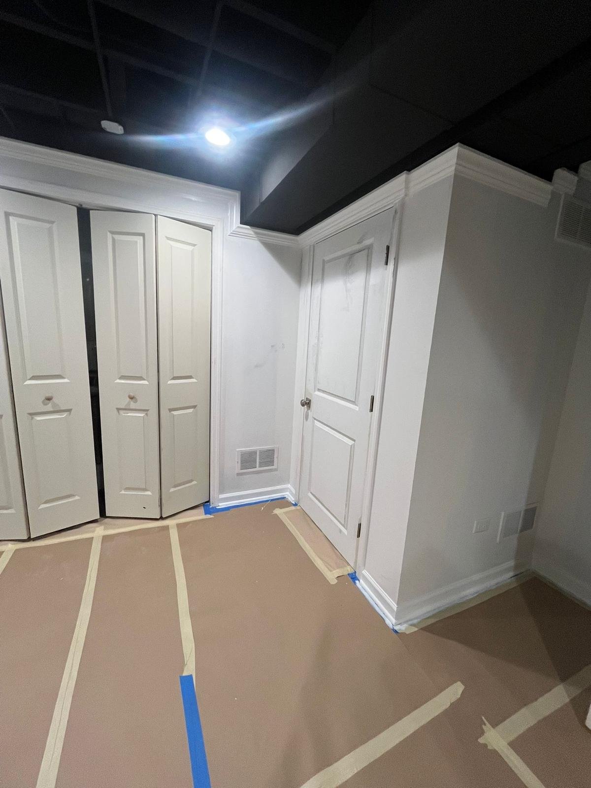 Room with white closet doors, white walls, and brown paper covering the floor, taped down with painter's tape, and a ceiling light turned on.