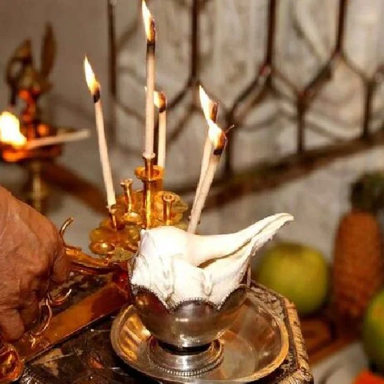 A white conch shell placed in a metallic bowl with lit incense sticks inserted into it, set on a decorative wooden surface.