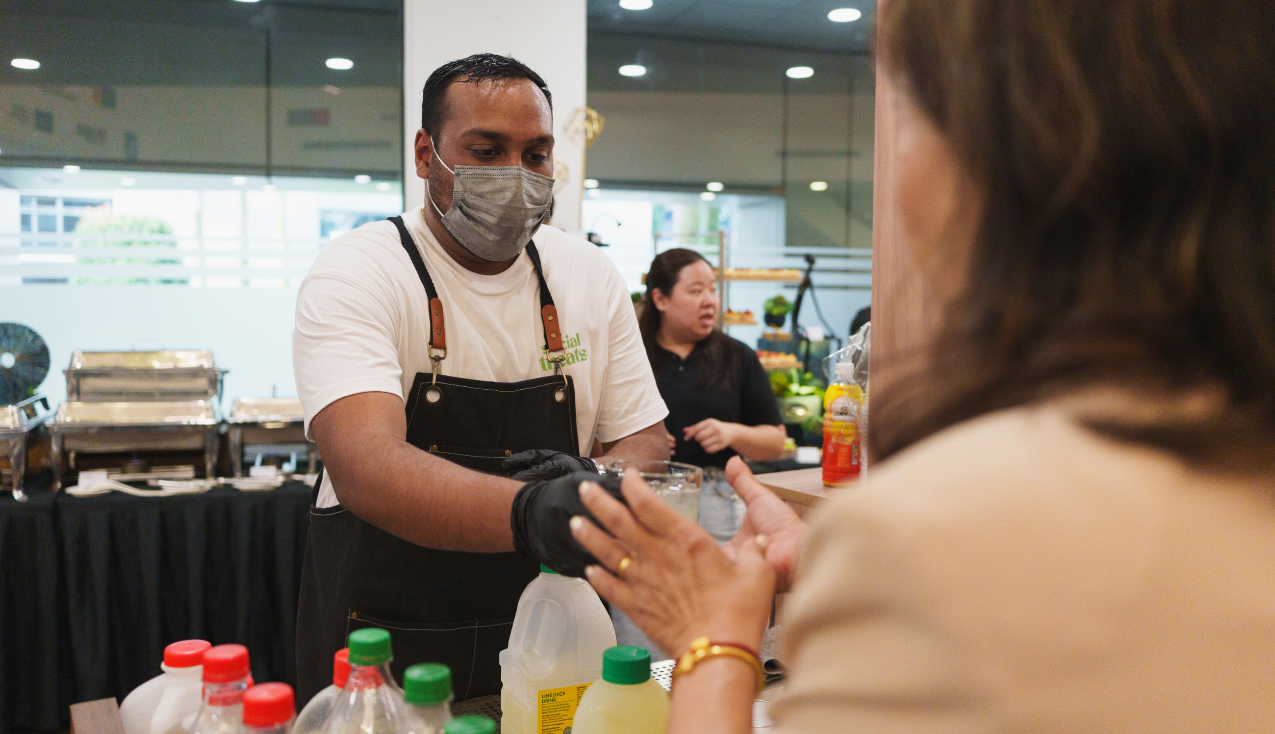 A man wearing a face mask and gloves serves a drink to a woman at a food or beverage stall. The man is dressed in a white t-shirt and black apron, with condiments and bottles on the counter in front of them.