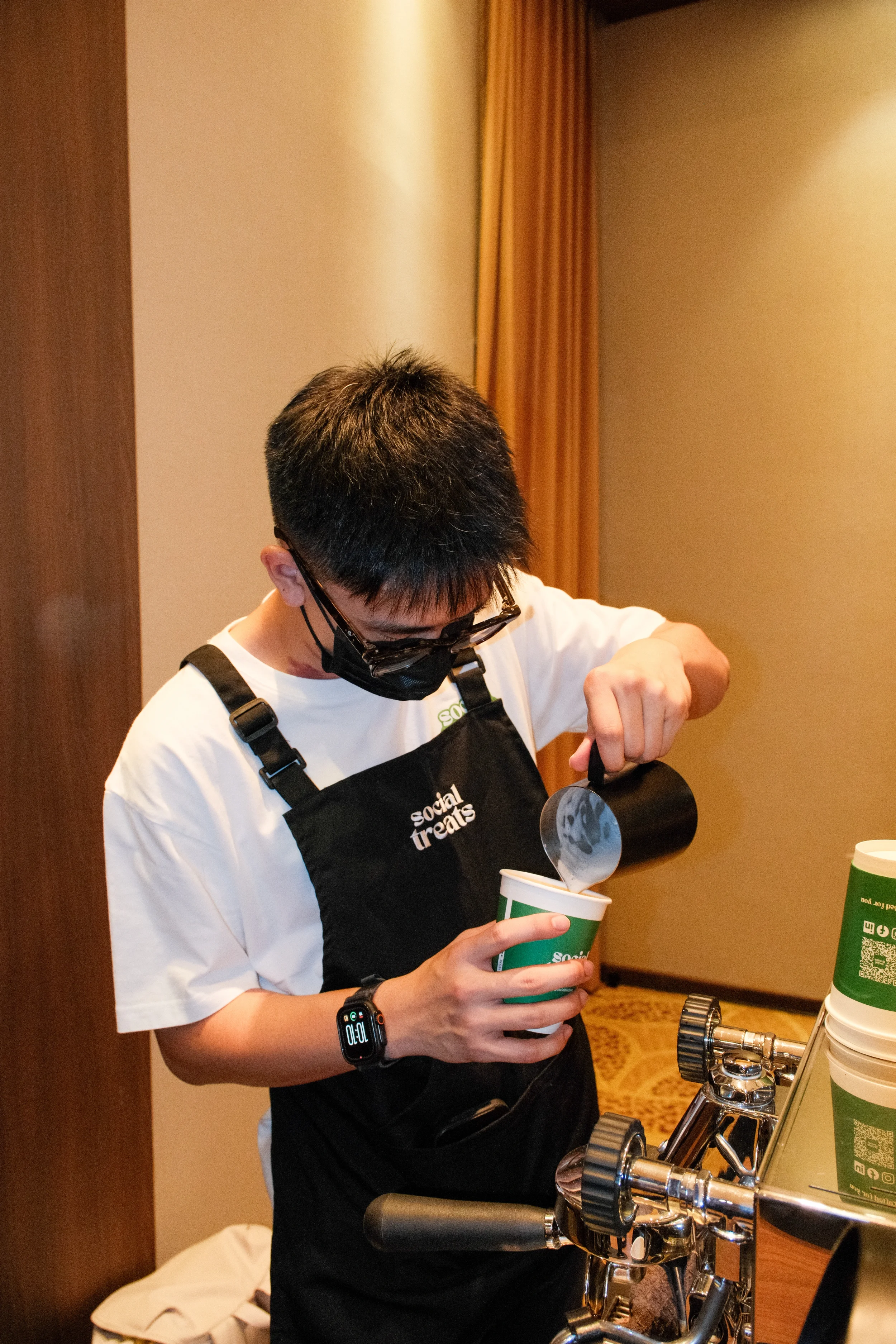 A person wearing glasses, a black face mask, and a black apron labeled 'social treats' is pouring a drink from a black pitcher into a green paper cup.
