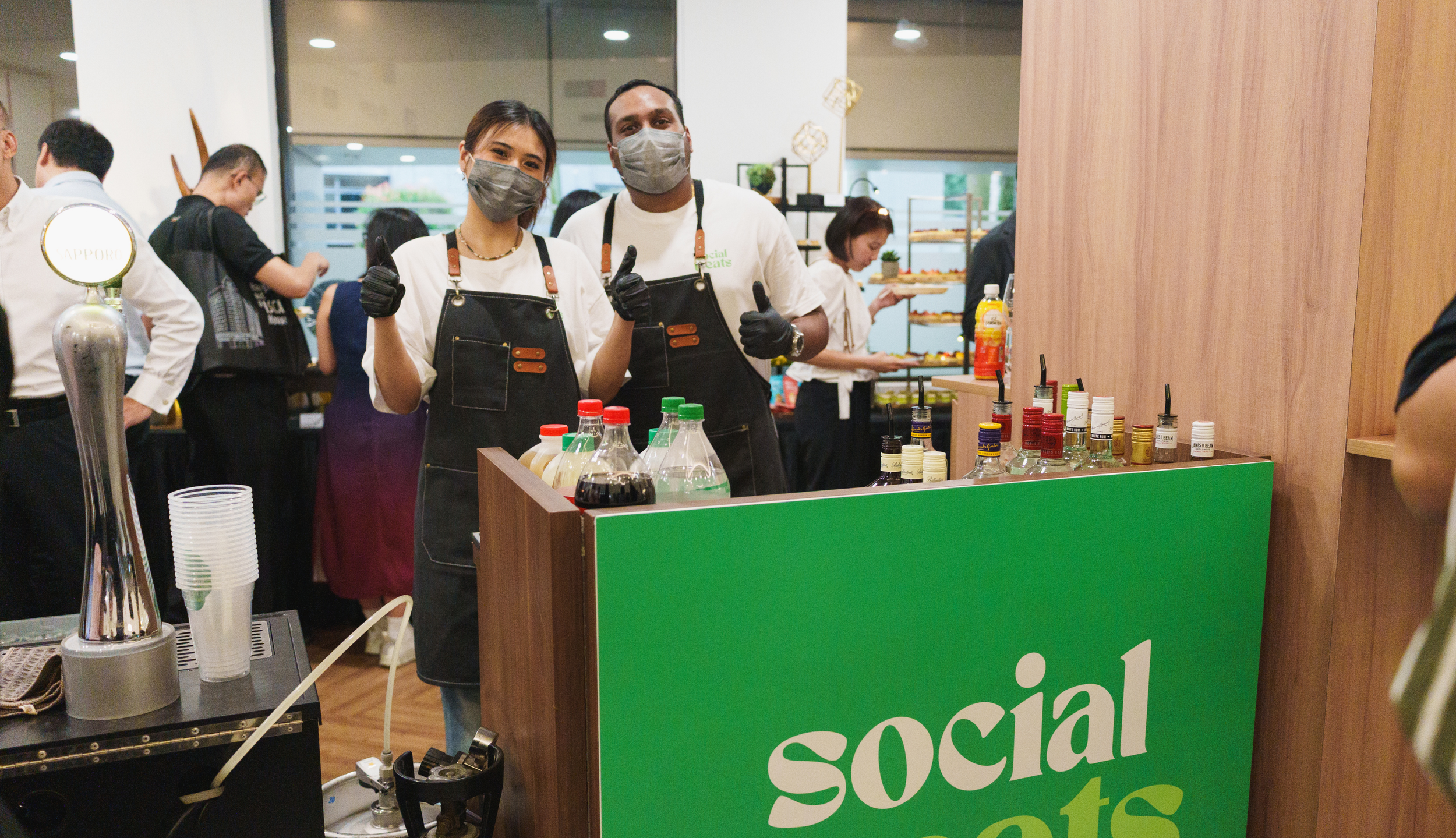 Two people wearing masks and aprons giving a thumbs-up behind a counter with condiments at a social eats event.