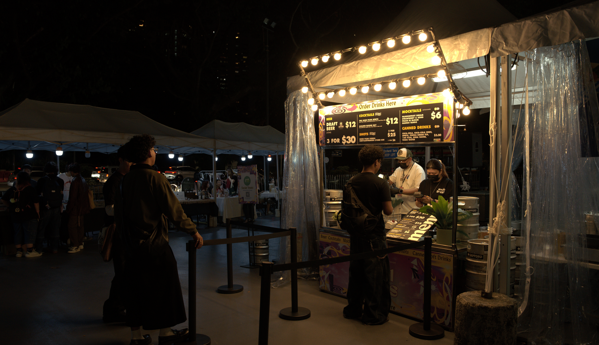 Night scene at a food or drink stand with illuminated sign and people ordering, under tents with other booths in the background.