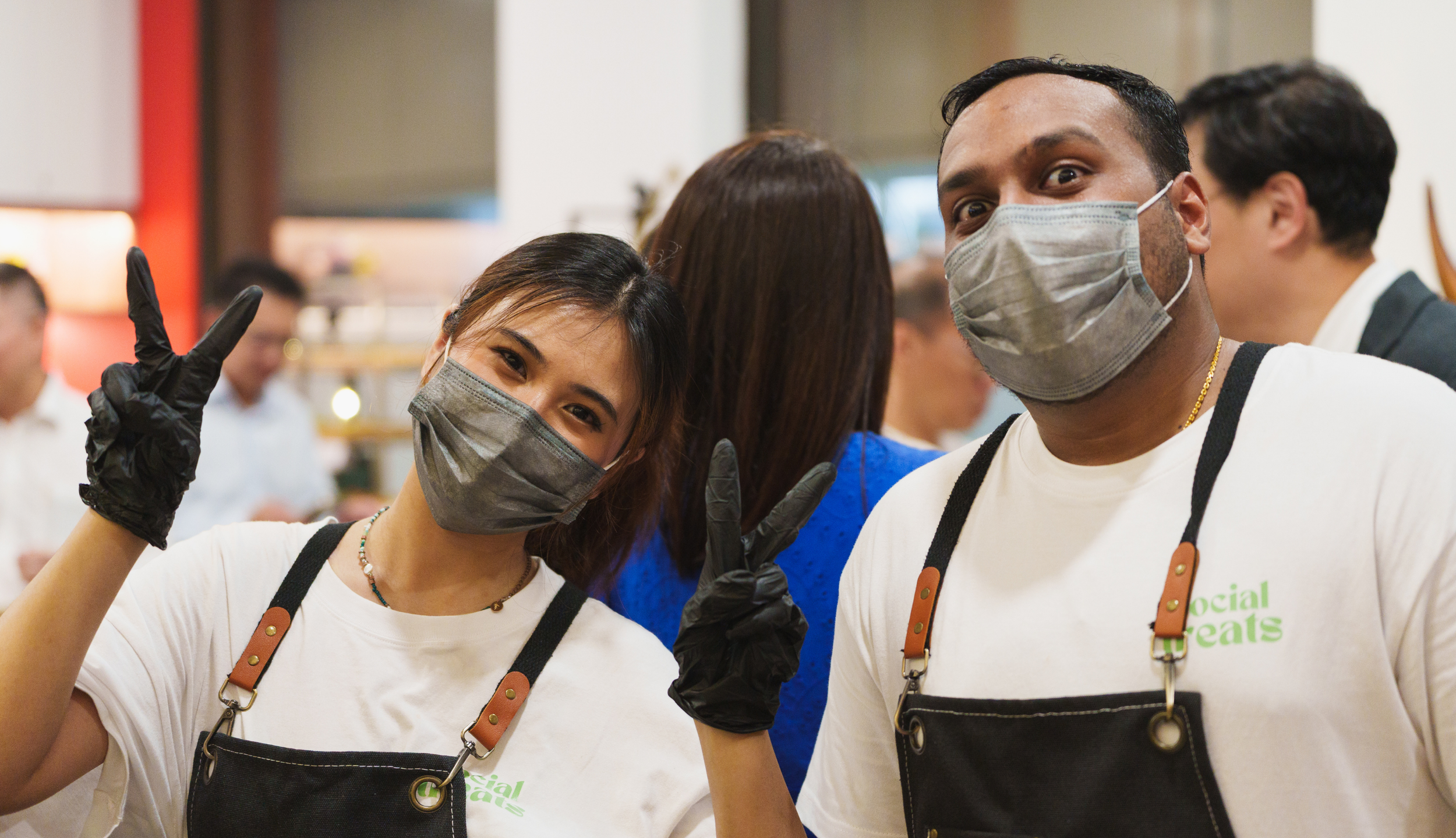 Two smiling volunteers in face masks, making peace signs at a social event.