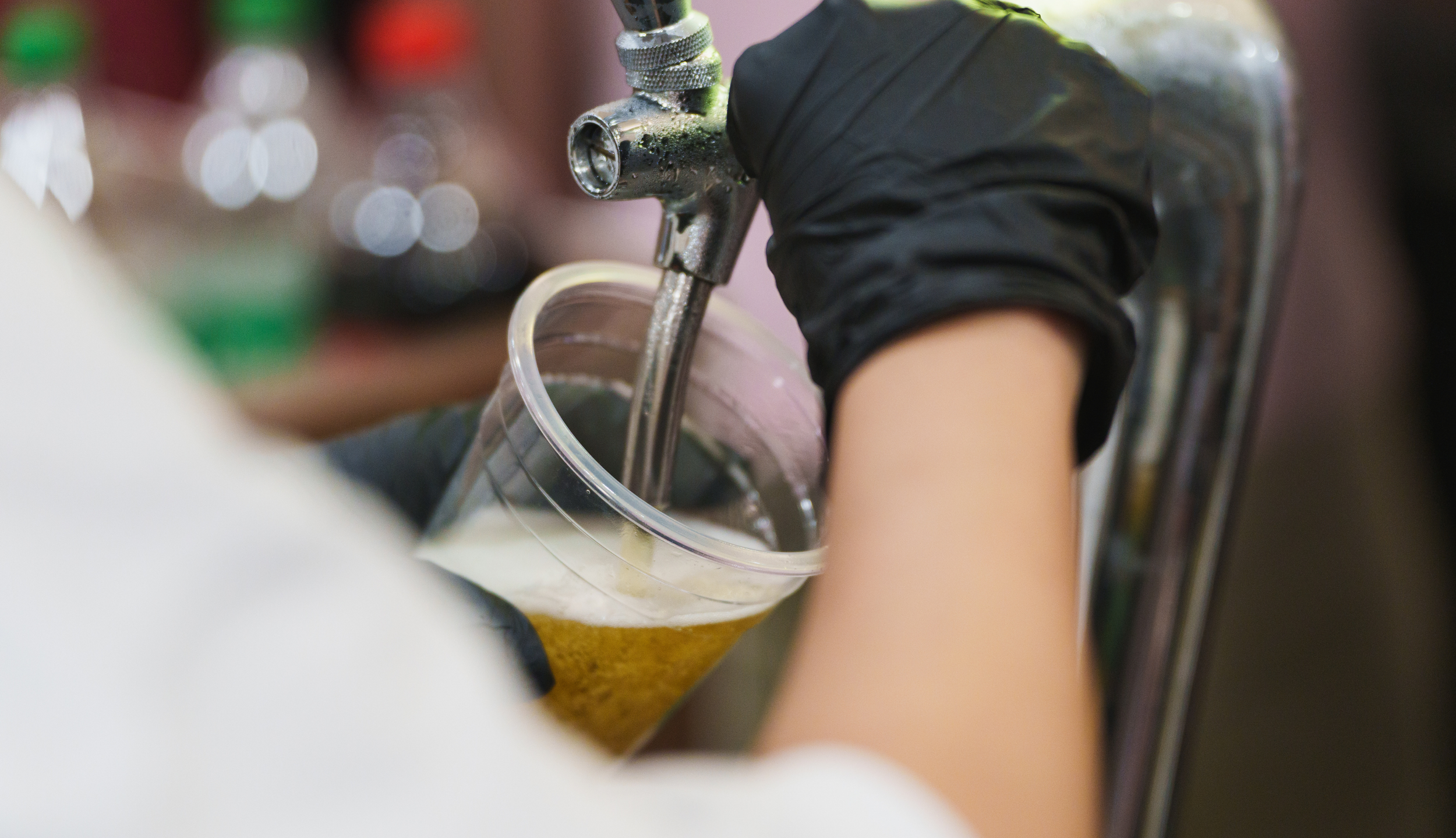 A person wearing a black glove filling a clear plastic cup with beer from a tap.