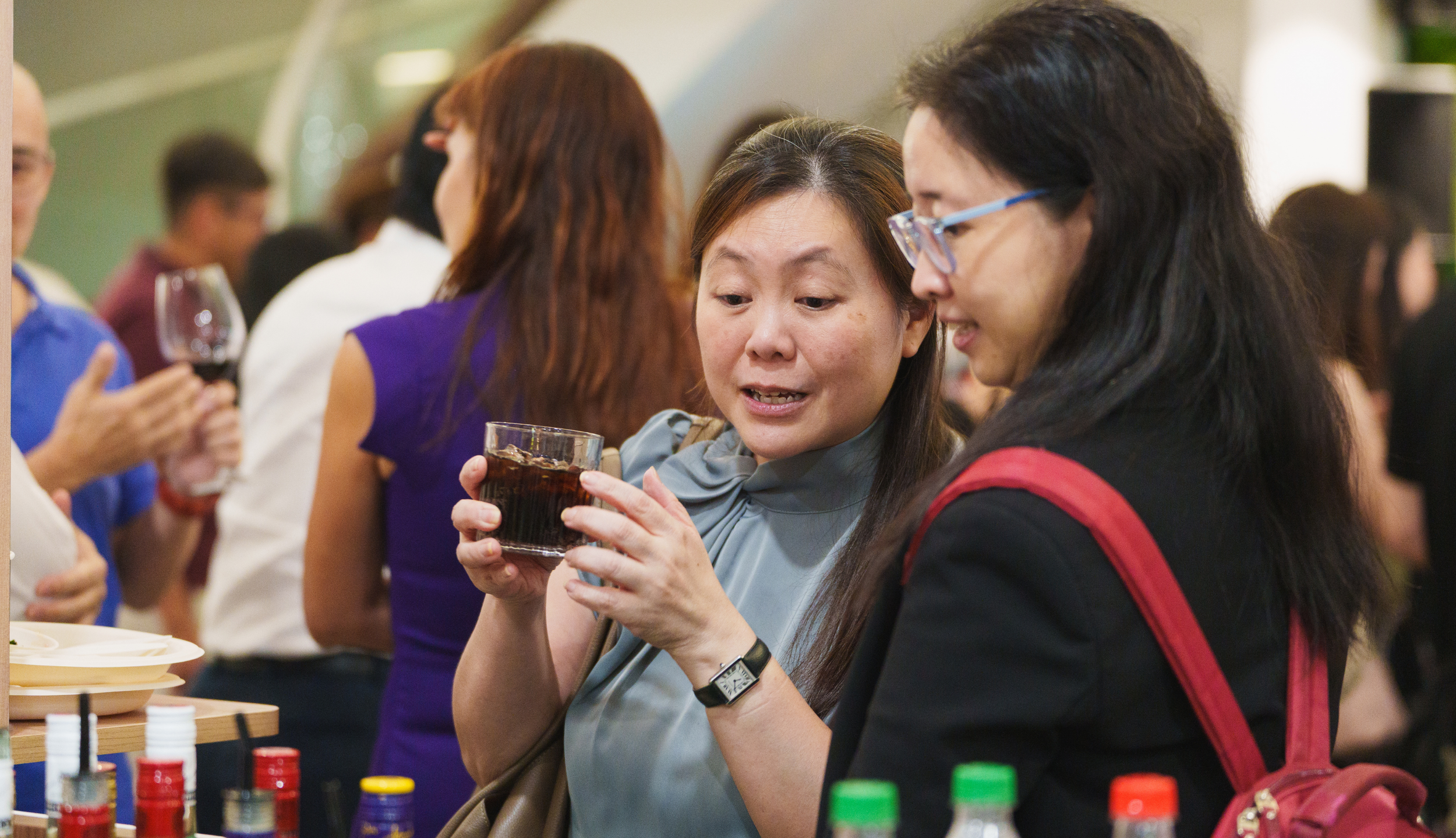 Two women at a social gathering, one holding a glass of cola and the other looking at it, with a crowd in the background.