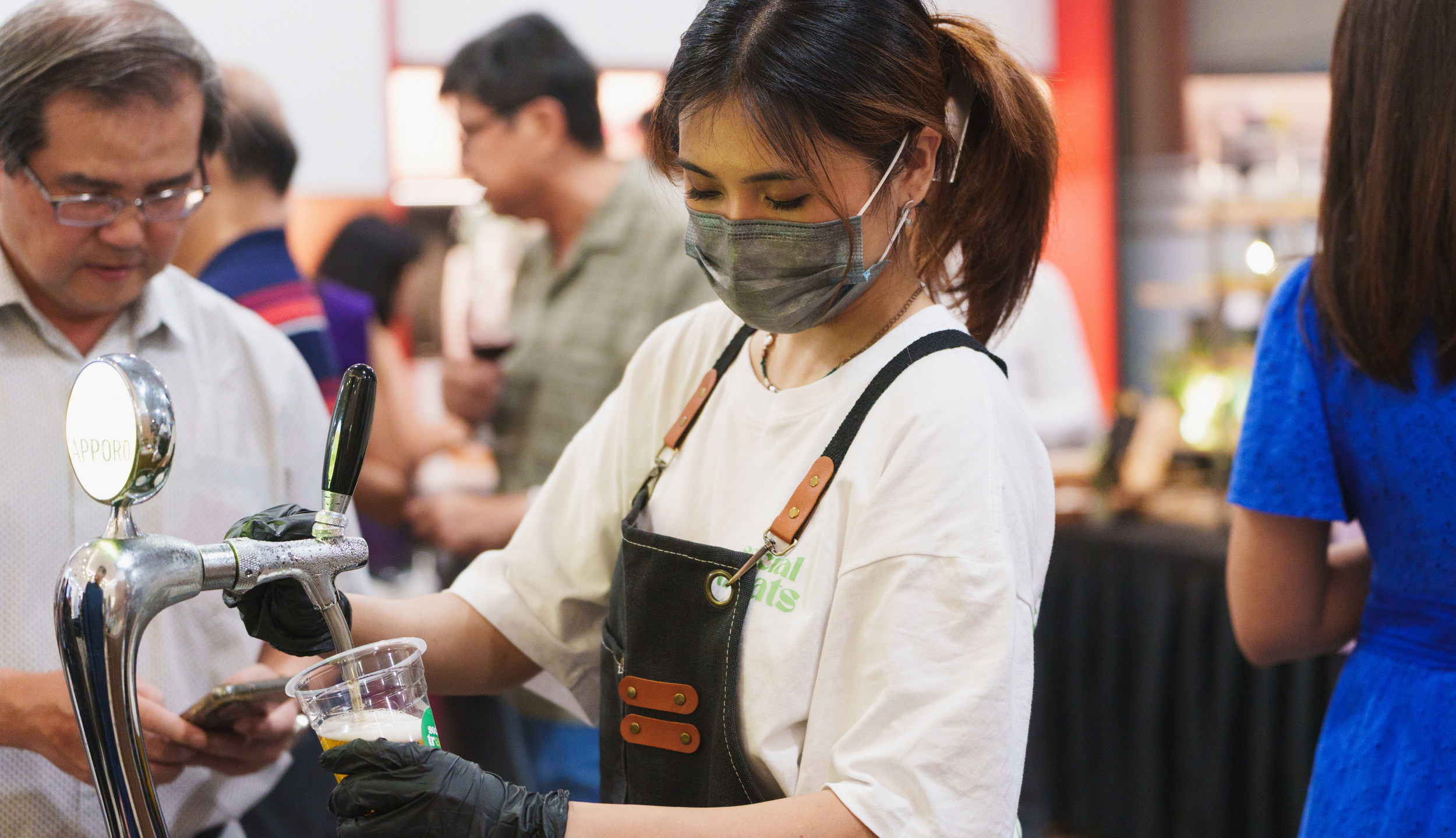 A woman with brown hair in a ponytail wearing a gray face mask, black gloves, and an apron is pouring a beverage from a tap into a plastic cup at a busy event or bar. Several people are in the background, some holding drinks, in an indoor setting.