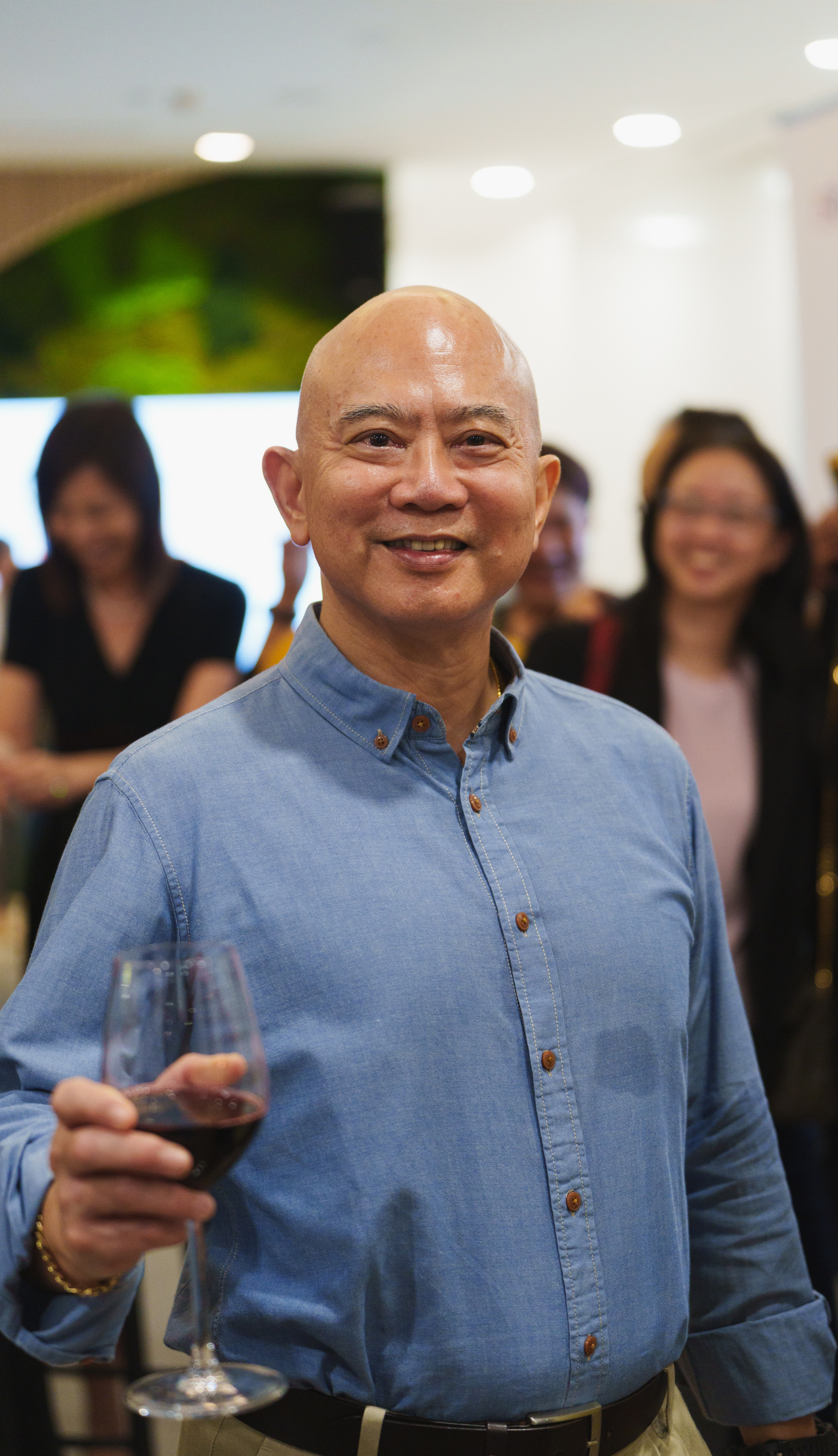 A smiling man with a shaved head holding a glass of red wine, wearing a blue button-up shirt, at a social gathering with blurred people in the background.