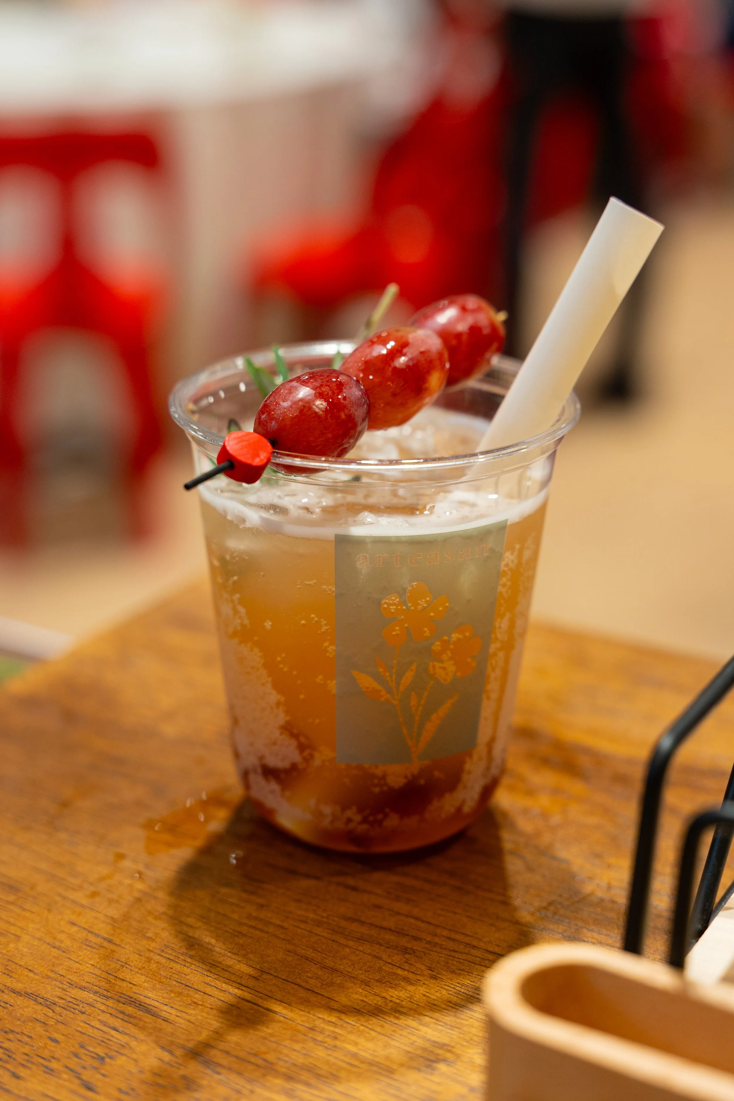 A clear plastic cup filled with a light brown beverage, garnished with three red grapes pierced by a black and red skewer, a white straw, and placed on a wooden surface.