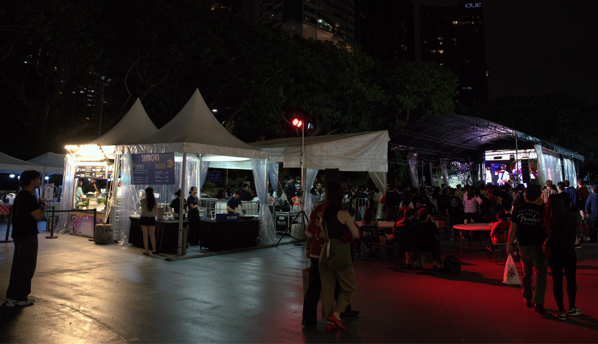 Night scene at an outdoor event with a stage, tents, and people. The stage has a band performing with purple and white lights. Several people are standing and sitting, enjoying the event. Some food stalls are visible under tents, and trees and tall b