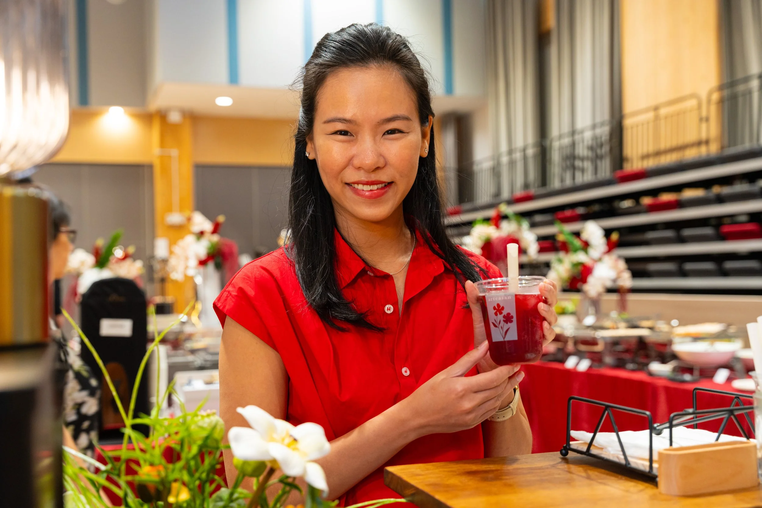 A smiling woman in a red shirt holding a clear plastic cup with a red drink and white straw in a decorated event space.