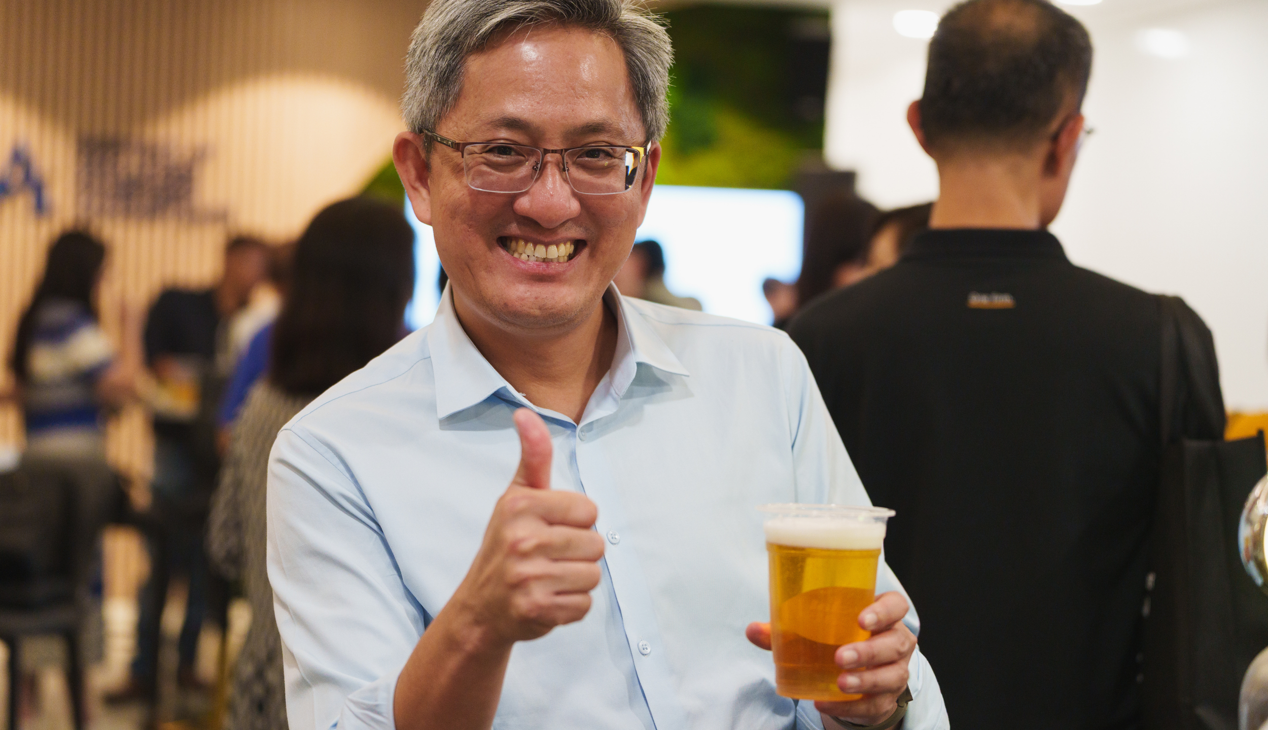 A smiling man with glasses in a white shirt holding a plastic cup of beer, giving a thumbs-up, at a social gathering.