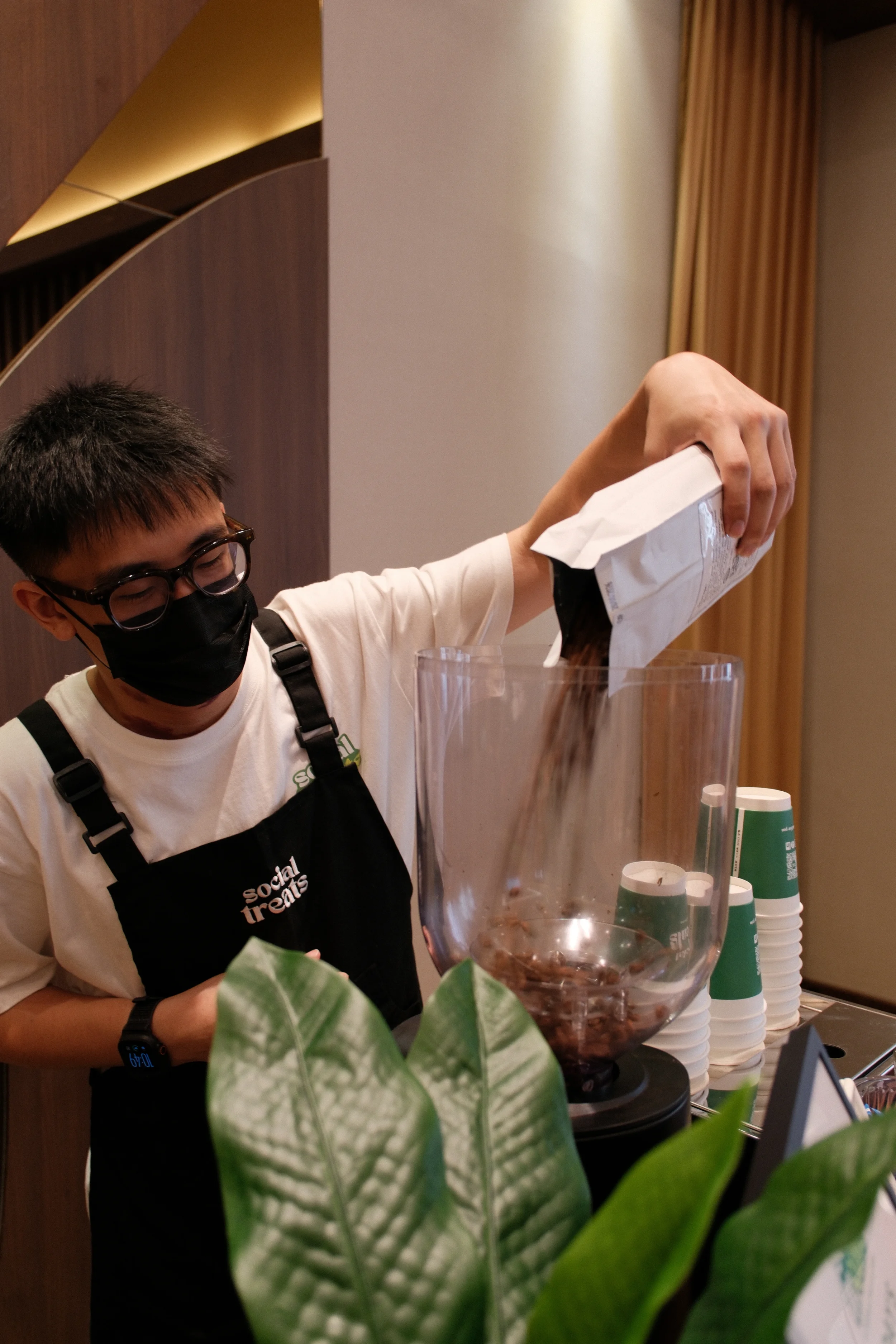 A person wearing glasses, a black face mask, and a black apron labeled 'Social Treats' is pouring chocolate from a white bag into a large transparent container, with green plants and stacked cups nearby.