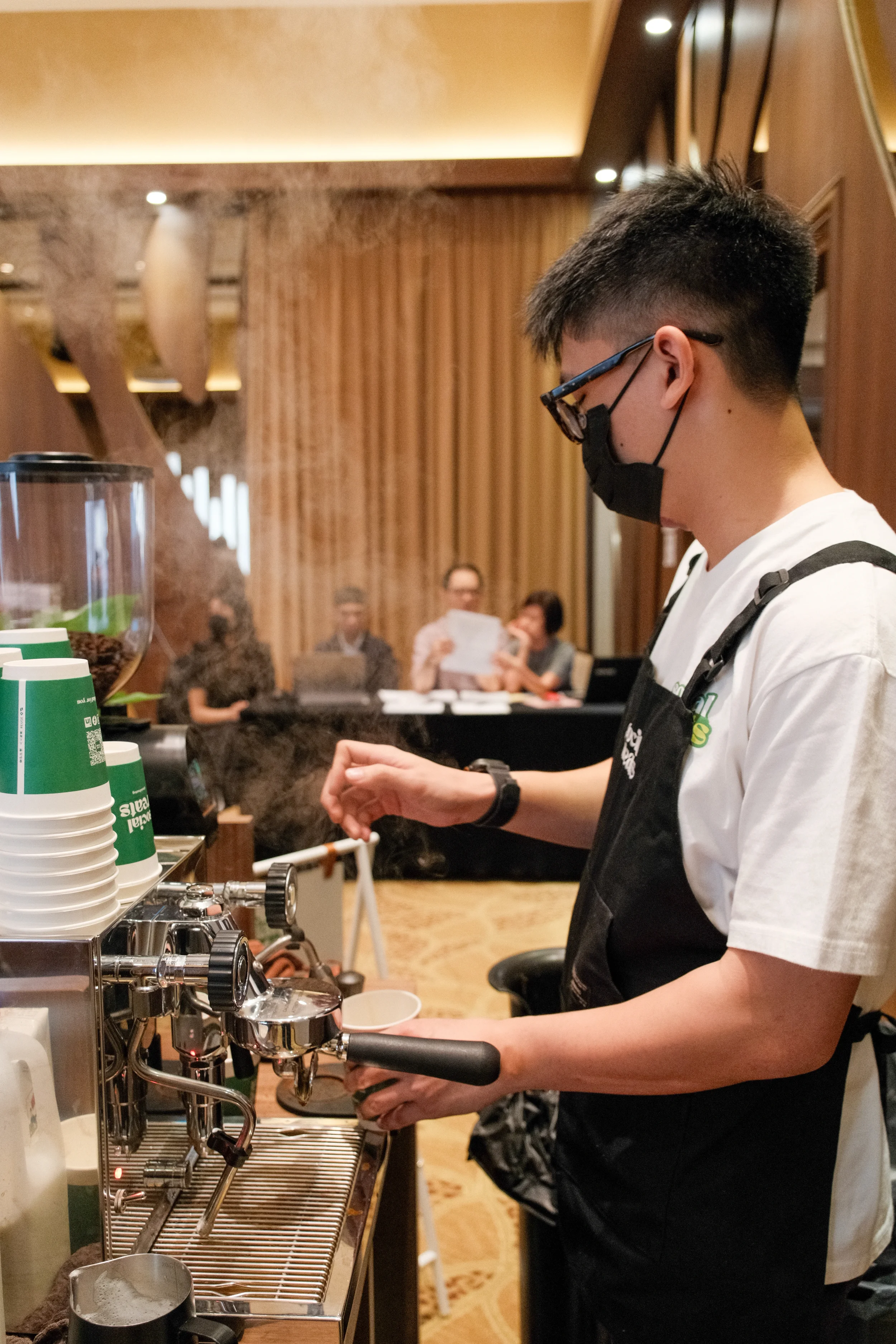 A barista wearing a black mask, glasses, and a white t-shirt preparing coffee behind a coffee machine in a cafe with wooden decor and several customers in the background.