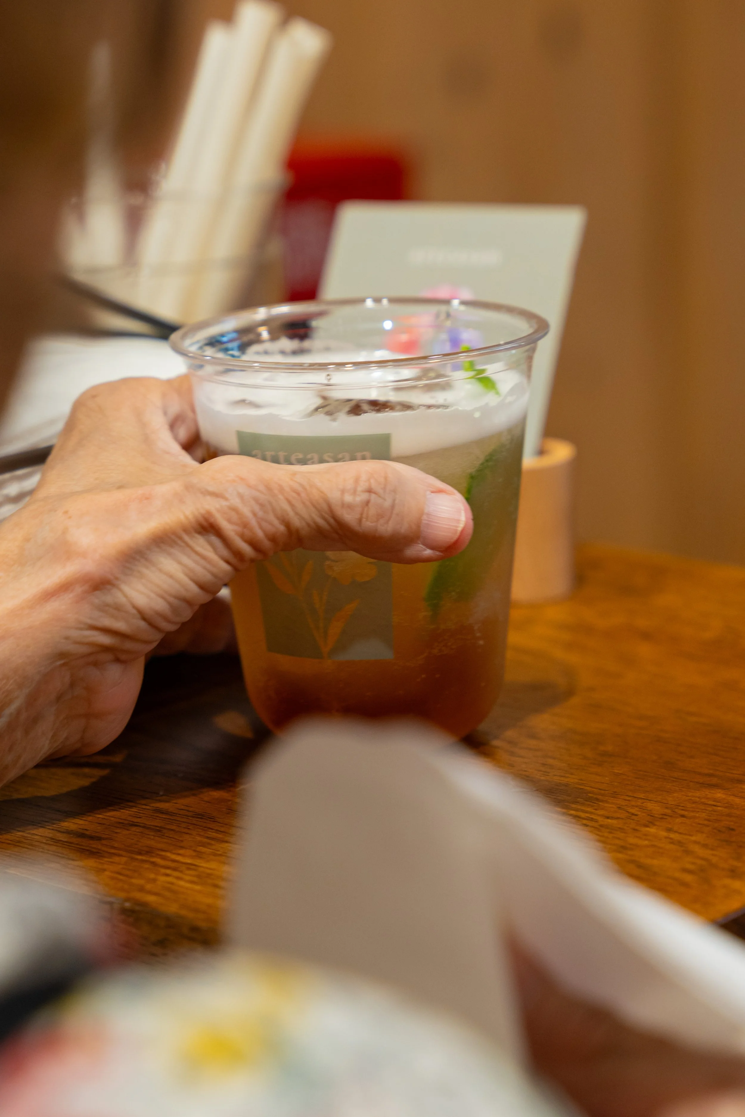 A person holding a plastic cup with a beverage, possibly iced tea, with lime slices inside, on a wooden table with other objects in the background.
