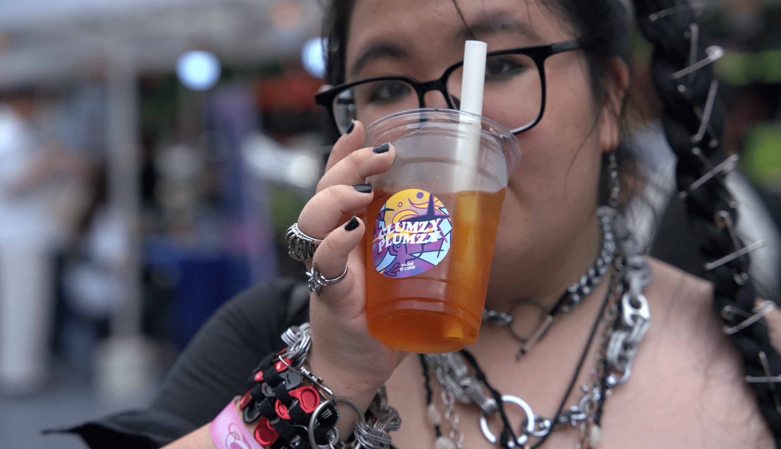 Person with dark hair, glasses, and multiple jewelry items is drinking a beverage from a clear plastic cup with a straw, outdoors.